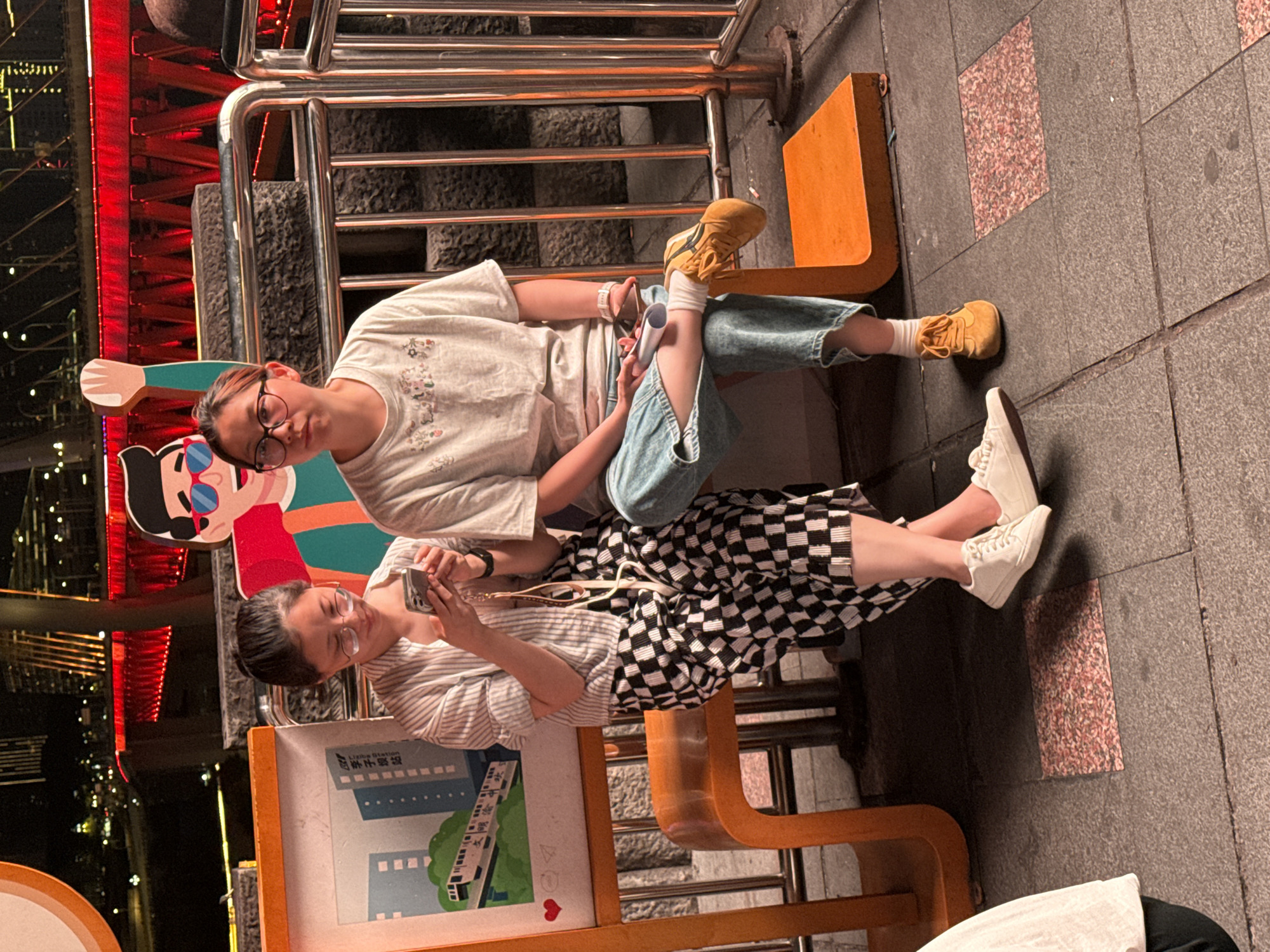 Two people sit on an orange-colored bench in Yuzhong District, China. One person wears glasses, a white t-shirt with images, blue jeans, and tan-colored shoes. The other person wears glasses, a vertically striped white and grey shirt, and a checkered black and white skirt. Both are using phones. The background shows a nighttime city scene with lights and reflections, a staircase, and a sign with an image of a building.
