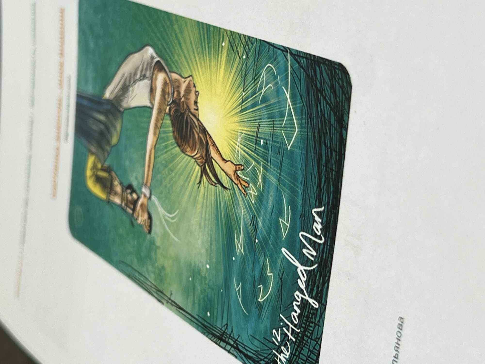The image shows a Tarot card, "The Hanged Man," lying on a white surface. The card features an illustration of a woman in a backbend pose, reaching towards a bright, yellow light source. She is wearing a white top and a dark, striped skirt. Her hair is brown and flowing. The background is a textured green with dark, vertical lines resembling trees. White, geometric symbols are scattered around the light source. The card's title, "The Hanged Man," and the number "12" are written in white script on the bottom right corner of the card. On the white surface below the card, a portion of the name "Lyanova" written in Cyrillic (probably льsянова) is visible. The location of Perm, Russia, is not indicated by the image.