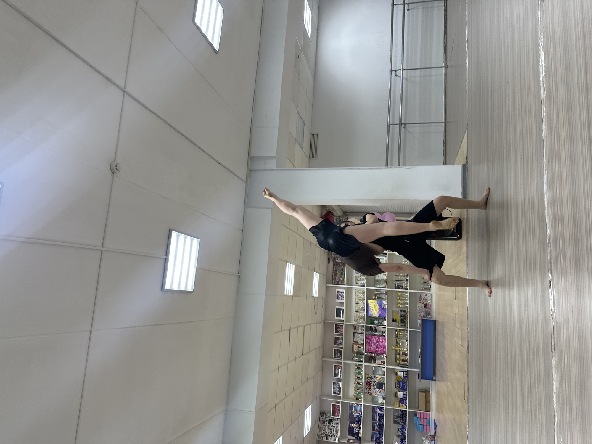 The image shows two people performing an acrobatic pose in what appears to be a gymnasium or dance studio. One person is lying on their back with their legs and arms extended upwards. The second person is balancing on top of the first person's feet and hands, also with their body extended. The room has a white ceiling with square lights. One wall has wooden floors and another wall has a display cabinet filled with trophies and memorabilia. The city and country are not discernable from the image.