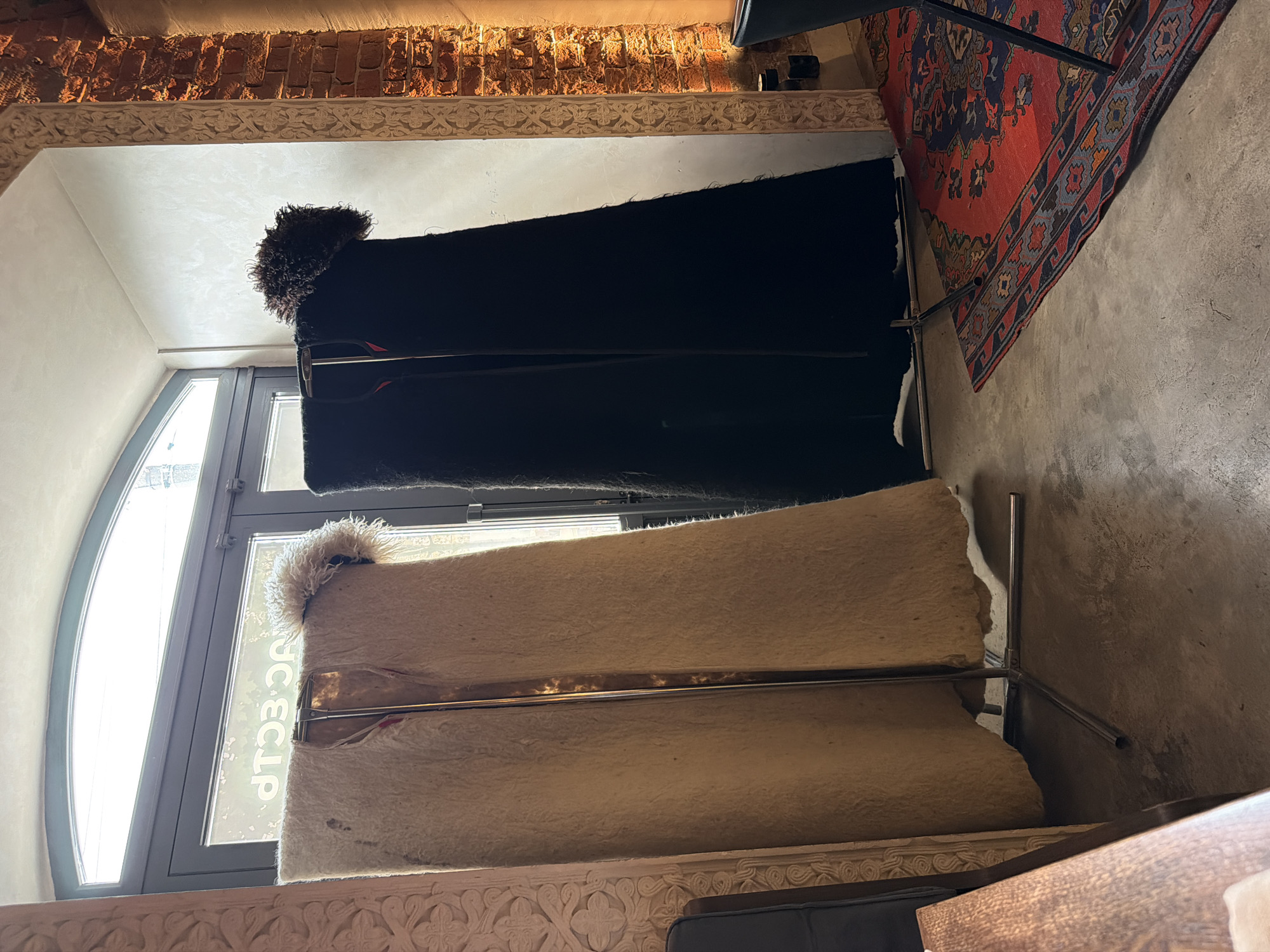 In an indoor setting with natural light filtering through a large arched window and a brick wall to the right, two tall, floor-standing garment racks display two large, draped garments. The garment on the left is a light-colored, possibly sheepskin, cape with a fluffy trim around the top. The garment on the right is a black cape, also with a dark, fluffy trim. Both garments hang open, revealing a glimpse of a red lining. The floor is concrete, and a red Persian rug with intricate patterns lies on the floor to the right of the racks. To the left, a carved wooden architectural element is visible. A portion of a dark chair and a wooden table are in the foreground on the left. The background shows a closed glass door with faint lettering on it and the brick wall illuminated by warm lighting.