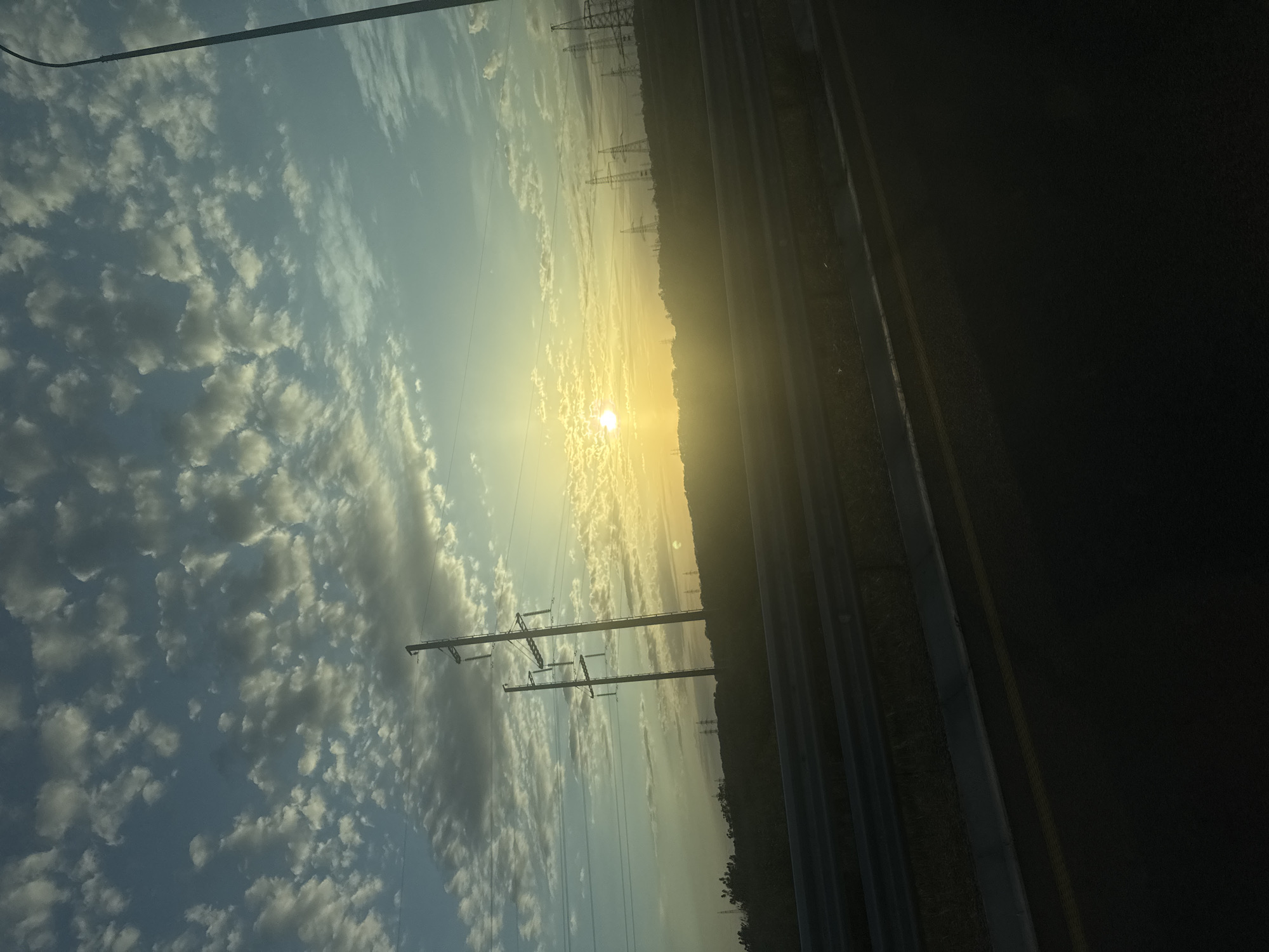 The image depicts a view of a landscape at what appears to be dawn or dusk, with a bright sun low on the horizon amidst a sky filled with scattered clouds. The perspective suggests it was taken from within or near a vehicle. Power lines and towers are visible in the middle ground, stretching across the scene. To the right of the image is a dark area, possibly a highway or road, marked with yellow lines. The scene suggests a rural or semi-rural setting. There is no evidence to confirm it is in Starotitarovskaya, Russia.