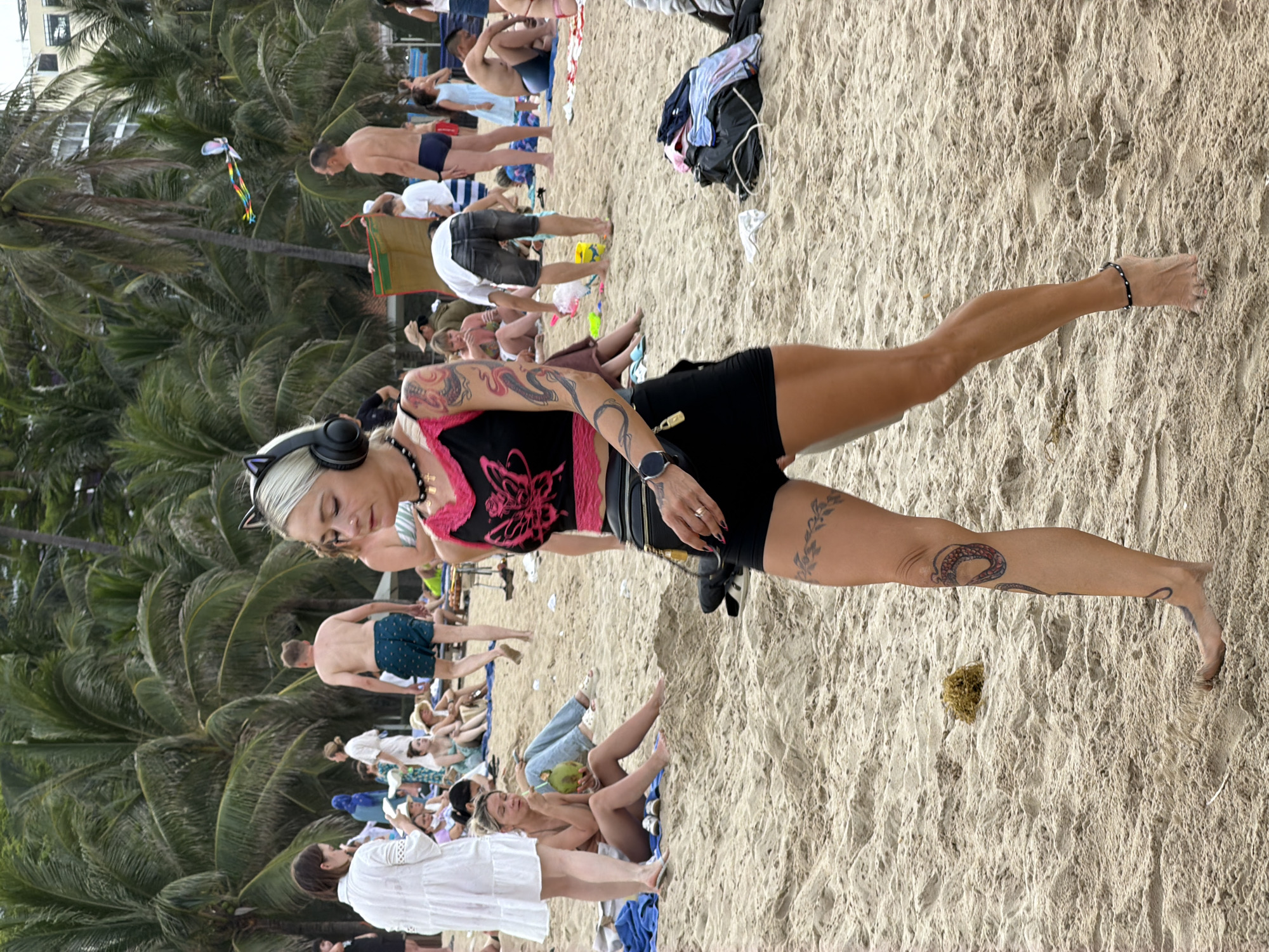 The image depicts a crowded beach scene in Nha Trang Ward, Vietnam. In the foreground, a woman with blonde hair, wearing cat-ear headphones, a black and pink crop top, and black shorts, walks across the sand. She has visible tattoos on her arms and legs, and is wearing a smartwatch and an anklet. In the background, numerous people are scattered across the beach, some sunbathing, others walking or sitting. Palm trees line the back of the beach, and some buildings are visible through the foliage. The sand is light brown and appears to be slightly wet in places. A kite can be seen flying in the sky above the palm trees. The overall atmosphere is that of a typical busy beach day.