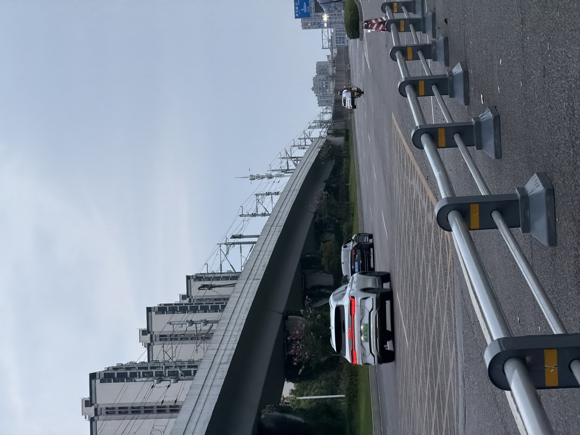 An overcast, daytime, eye-level, medium shot captures a road with traffic, flanked by a raised overpass and apartment buildings on the left, and a metal guardrail and traffic cones on the right. The road is asphalt with yellow and white lane markings. In the foreground, a grey metal guardrail with yellow reflective markers runs along the right side of the road. Beyond the guardrail, a few small bits of debris are visible on the asphalt. In the middle ground, a white SUV with its taillights illuminated travels in the left lane of the road, followed by a dark sedan. Further down the road, a person rides a scooter in the same direction. A few indistinct buildings and trees are visible in the distance. On the left side of the road, a concrete overpass, supported by substantial pillars, spans the scene. Electrical wires for what appears to be a train or elevated transit system are strung across the top of the overpass. Behind the overpass, a row of tall, modern apartment buildings with dark window frames and white facades are visible against the grey sky. The city of Jiangjin, China, is the implied location of this urban scene.