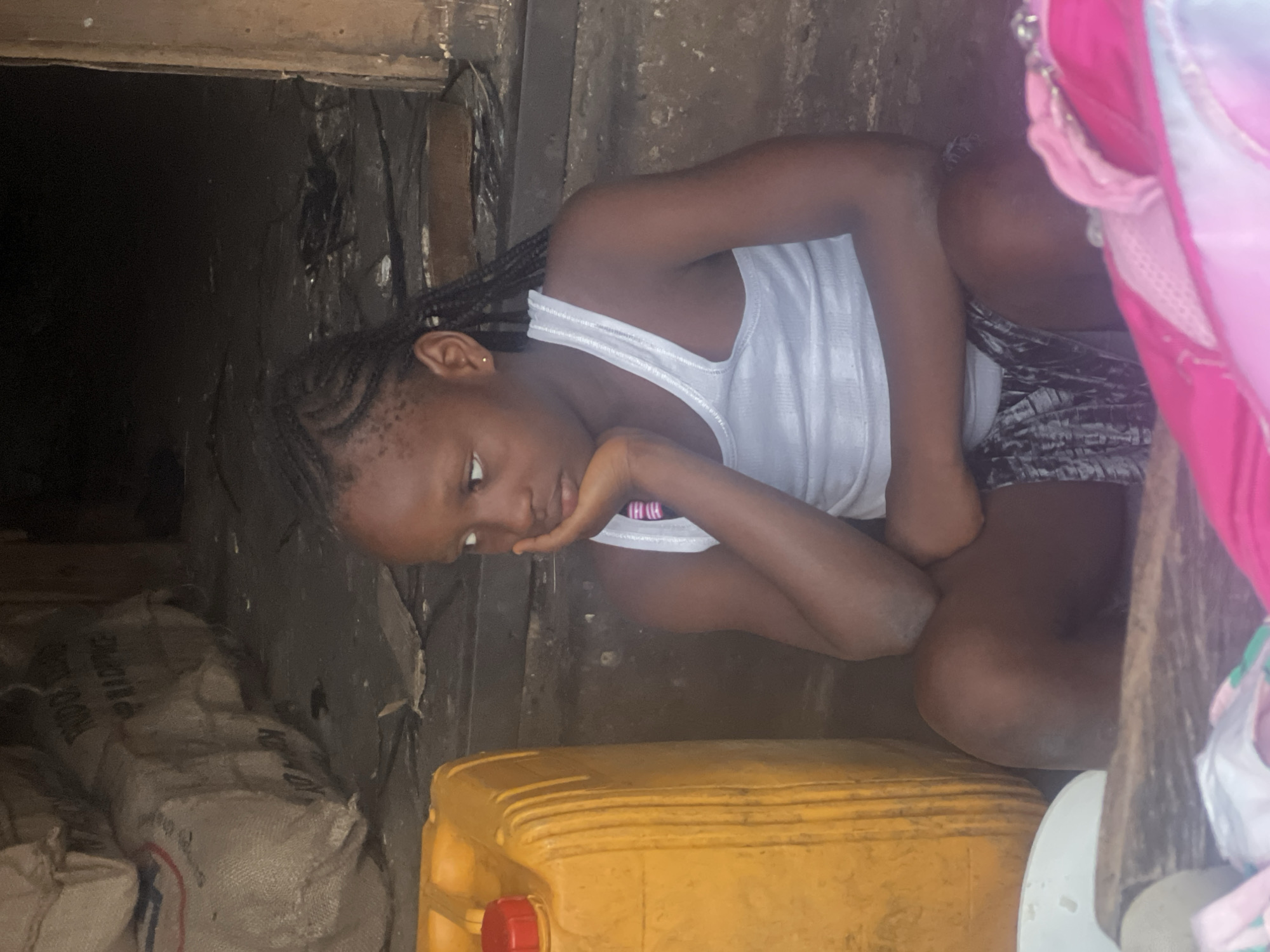 A young girl is depicted sitting in a dimly lit, enclosed space, likely in Nkpor, Nigeria. She appears to be of African descent, with braided hair, and is wearing a white tank top and dark patterned shorts. Her posture is somewhat crouched, with her knees drawn up, and she rests her chin on her left hand, looking downwards with a pensive or contemplative expression.
The setting features rough, dark walls, possibly made of wood or concrete, suggesting a simple or utilitarian interior. To the left of the frame, several large, light-colored sacks are stacked, some with visible creases. Partially visible text on one sack indicates "PRODUCED" or a similar word. In the foreground, a prominent bright yellow plastic jerry can with a red cap sits on the ground. To the far right, partially visible pink and white fabric or clothing adds a touch of color. The overall atmosphere is quiet, with the dim lighting suggesting an indoor environment with limited natural light.