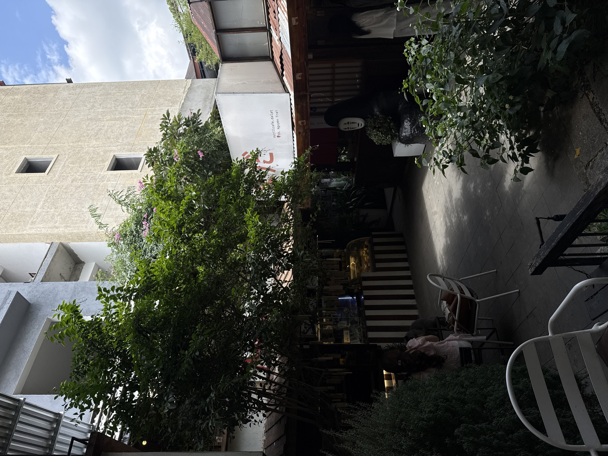 The image depicts the outdoor seating area of a cafe, likely located in Lâm Viên - Đà Lạt, Vietnam. The scene is characterized by a mix of natural elements and urban structures. In the foreground, several white metal chairs are arranged on a stone-tiled patio, some partially obscured by lush green bushes. A woman in a light pink dress is seated at a table in the midground, her back to the camera, with a handbag placed on an adjacent chair.
Behind the seating area, a row of businesses or shops is visible, featuring a striped facade with a glass display case showcasing pastries. A large, dark statue with a white, mask-like face, reminiscent of the character No-Face from Spirited Away, stands prominently in front of one of the establishments. Above this, a large white sign displays "#stillcafe.dalat" and "59. Nguyen Trai," indicating the cafe's name and address.
The background is dominated by a multi-story building with a grey, unfinished facade, suggesting ongoing construction or renovation. Another building with a textured, beige exterior and two dark rectangular windows is also visible. A vibrant green tree with abundant foliage and pink blossoms arches over the scene, providing shade. The sky above is a mix of blue and white clouds. The overall impression is that of a charming, albeit somewhat eclectic, urban cafe setting infused with greenery.