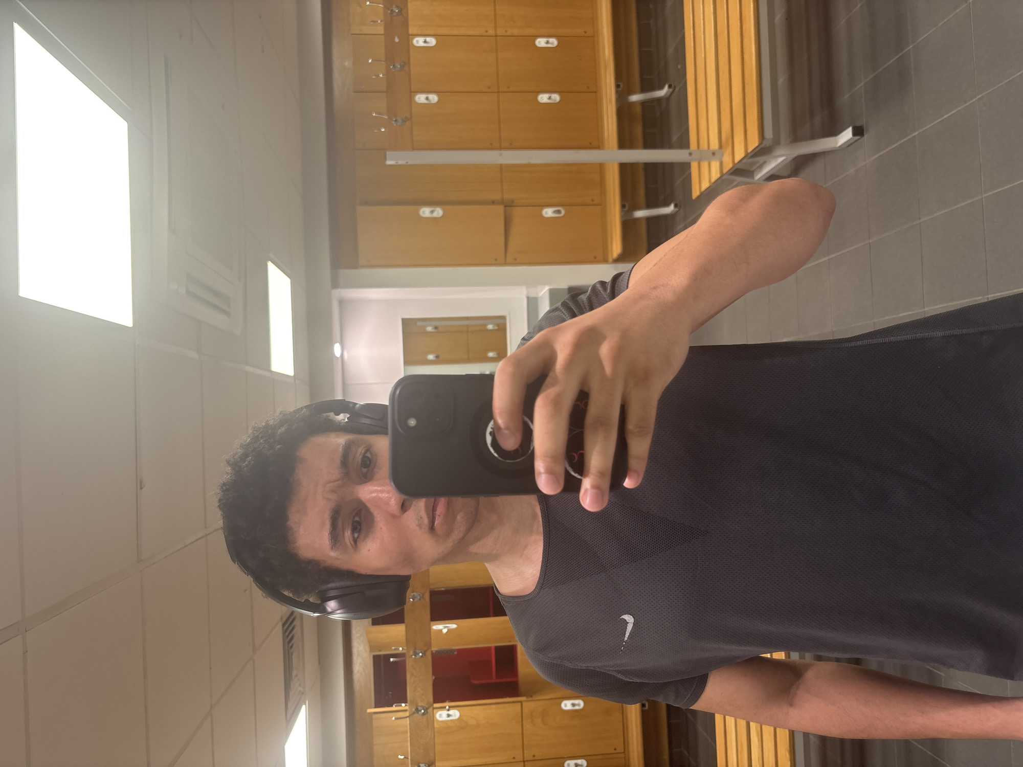 A person is taking a selfie in what appears to be a locker room. The individual is wearing headphones and a gray Nike t-shirt. The background consists of lockers and a bench. The location is likely a gym or sports facility, but there are no details that identify this as being in London.