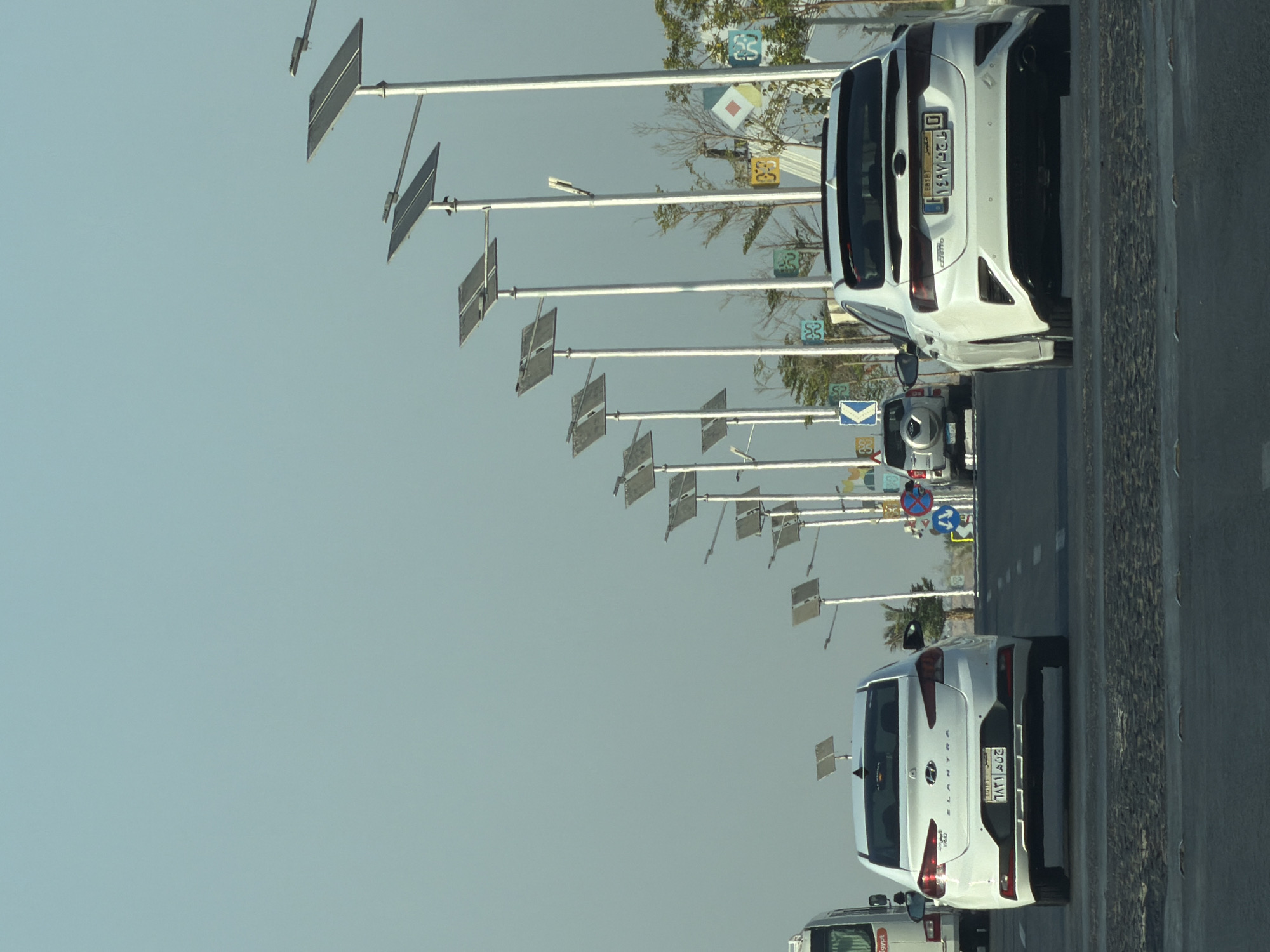 The image captures a view of a multi-lane road in Sharm El Sheikh, Egypt, under a clear, pale blue sky. A procession of solar-powered streetlights with large rectangular panels lines the right side of the road, extending into the distance. Several vehicles are visible on the road. In the foreground, the rear of a white sedan, possibly a Kia Cerato, is prominent, displaying an Egyptian license plate. To its left, another white sedan, identified as a Hyundai Elantra, is also seen from the rear, with its own Egyptian license plate. Further ahead, a silver SUV is parked or moving slowly, and a white bus is partially visible on the far left. Decorative square panels in various colors, featuring geometric patterns, are mounted on poles along the roadside, interspersed with vegetation. Traffic signs, including directional arrows, are also present. The road surface appears to be asphalt, with visible lane markings. The scene suggests an urban or suburban setting within Sharm El Sheikh, characterized by modern infrastructure and a generally clear and bright atmosphere.