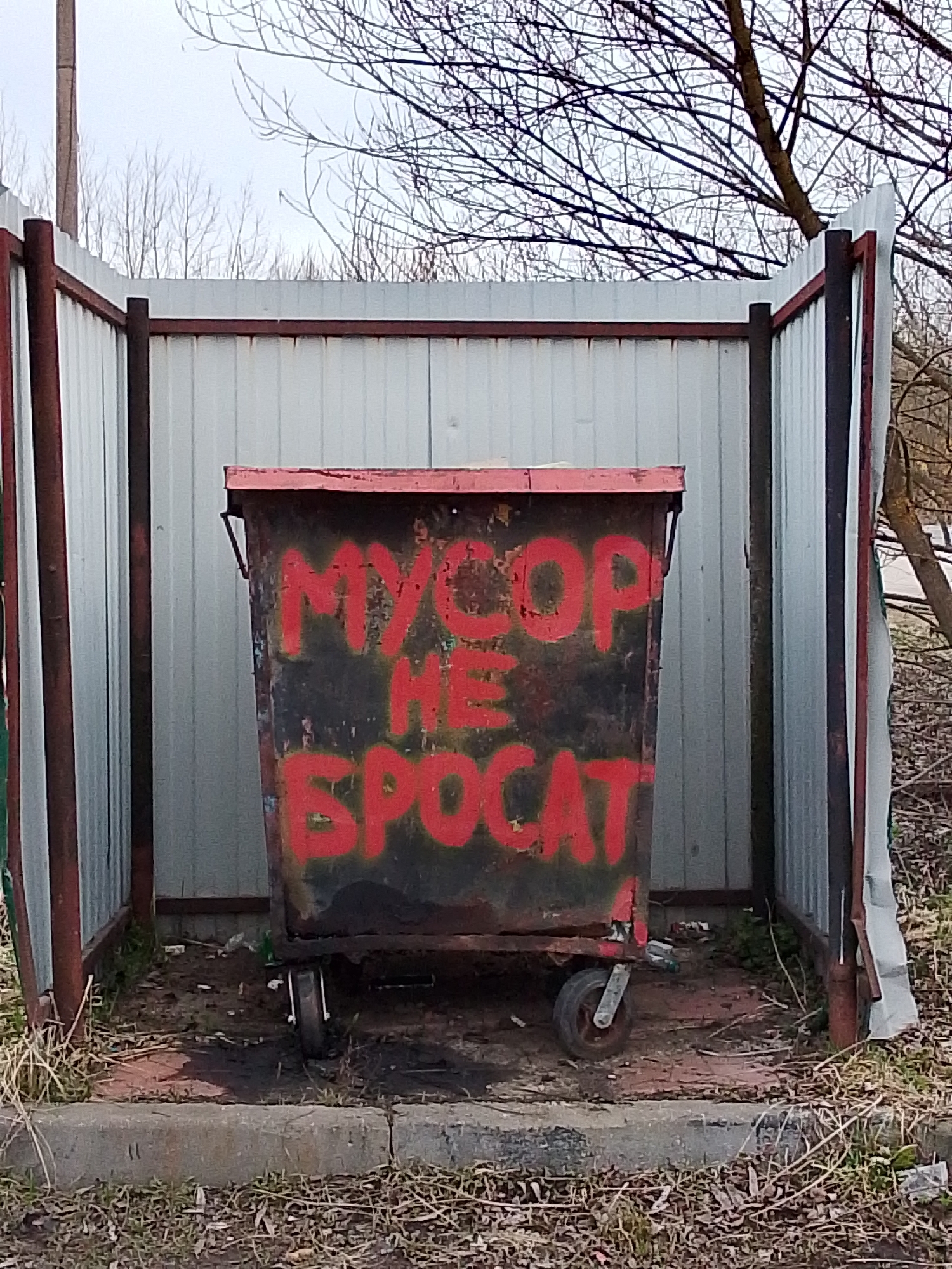A medium, centered shot reveals a trash container within a fenced enclosure located in the city of Снопок Старый, Russia. The trash container, predominantly black, has the phrase "MYCOP HE SPOCAT" spray-painted in red. The container is equipped with wheels and appears to be in a state of disrepair. The enclosure surrounding the trash container features corrugated metal sheets and dark metal posts, showing signs of rust and weathering. The ground inside the enclosure is littered with debris. Outside the enclosure, the environment includes dry grass, bare branches, and a concrete curb. The sky above is overcast.
