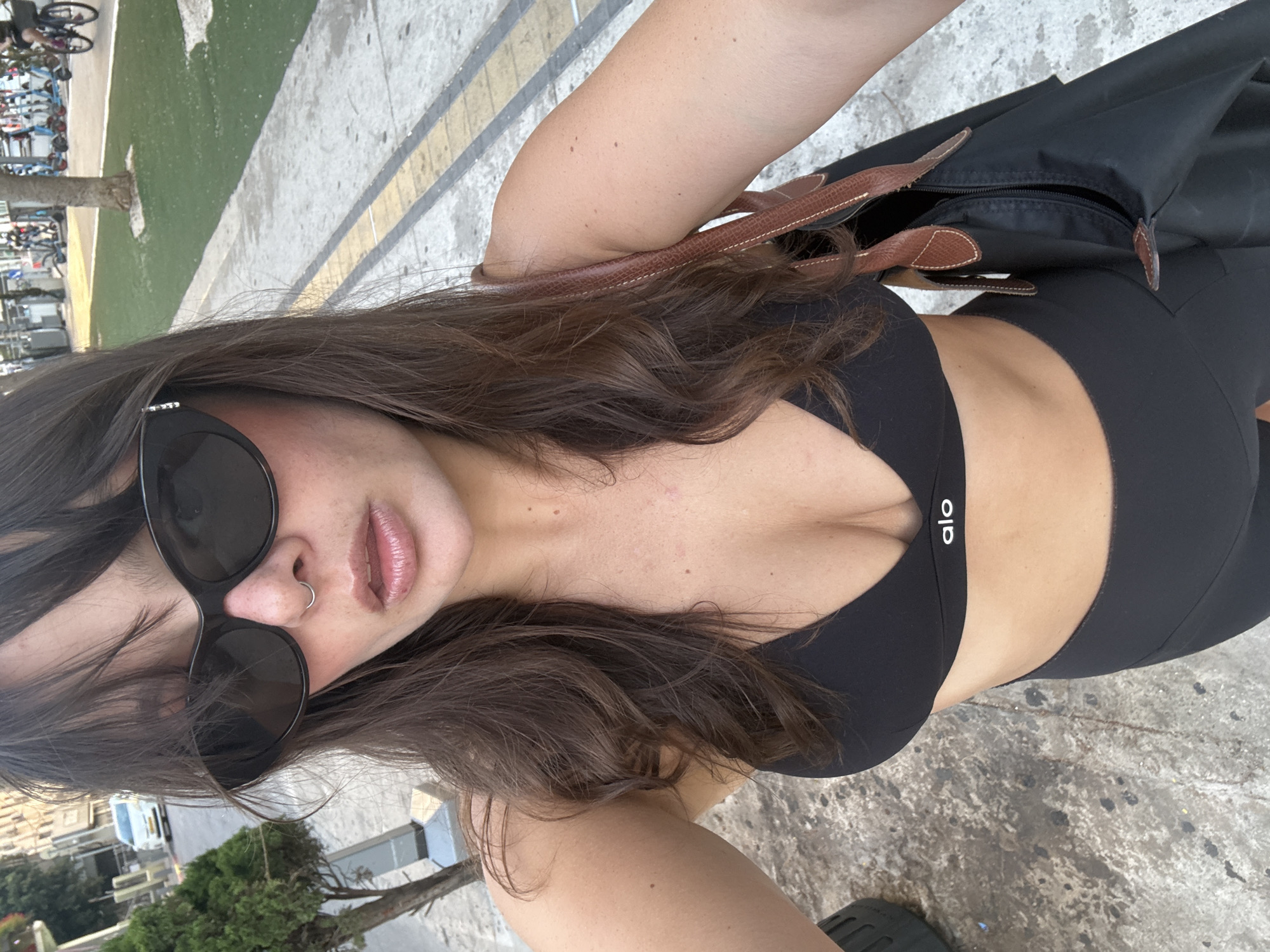 A woman with dark, wavy hair and sunglasses is taking a selfie outdoors in what appears to be a city environment. She is wearing a black sports bra with the "alo" logo visible and black leggings. A brown leather-strapped black bag is slung over her right shoulder. The background shows a sidewalk, a street with a white car, a tree, and a grassy area with parked electric scooters and bicycles, suggesting a public space within a city. The woman has a septum ring and a visible nose ring. The overall lighting suggests it is daytime. The scene is likely in Tel Aviv, Israel, given the style of the street furniture, vehicles, and the presence of electric scooters, which are common in the city.