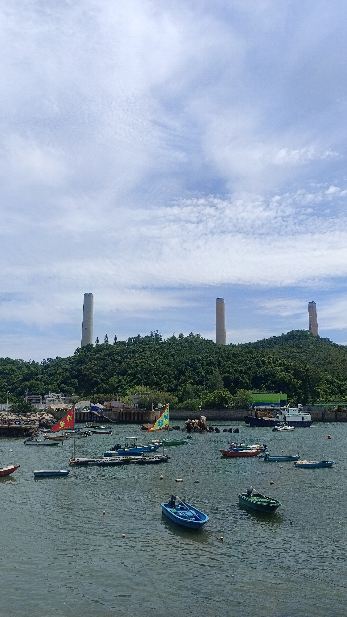 The image depicts a coastal scene with several boats moored in the water. In the background, a lush green, tree-covered hill rises, topped by three large, cylindrical smokestacks. The sky above is partly cloudy with wispy cirrus clouds. On the hillside, there are some buildings and structures, and a few red and yellow banners are visible, one with Chinese characters. Several smaller boats, including a blue dinghy and a green boat, are floating in the foreground. A larger white and blue boat is docked in the mid-ground, along with a small tugboat. The overall scene suggests a harbor or bay area. This location is likely the Lamma Power Station on Lamma Island, Hong Kong, China, known for its distinctive cooling towers and scenic coastal environment.