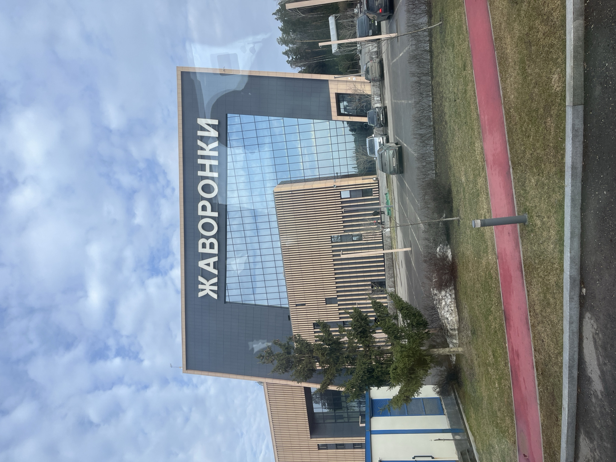 The image shows the exterior of a building with the Russian word "ЖАВОРОНКИ" vertically displayed on its facade. The building features a modern design with a glass curtain wall reflecting the sky, along with sections of horizontal wooden slat cladding. A row of parked cars is visible along a road adjacent to the building. A grassy area with a red running track runs alongside the road. There are trees present near the building. Based on the name on the building and the request, the location is likely in the city of Истра Гоу Доц, Russia.
