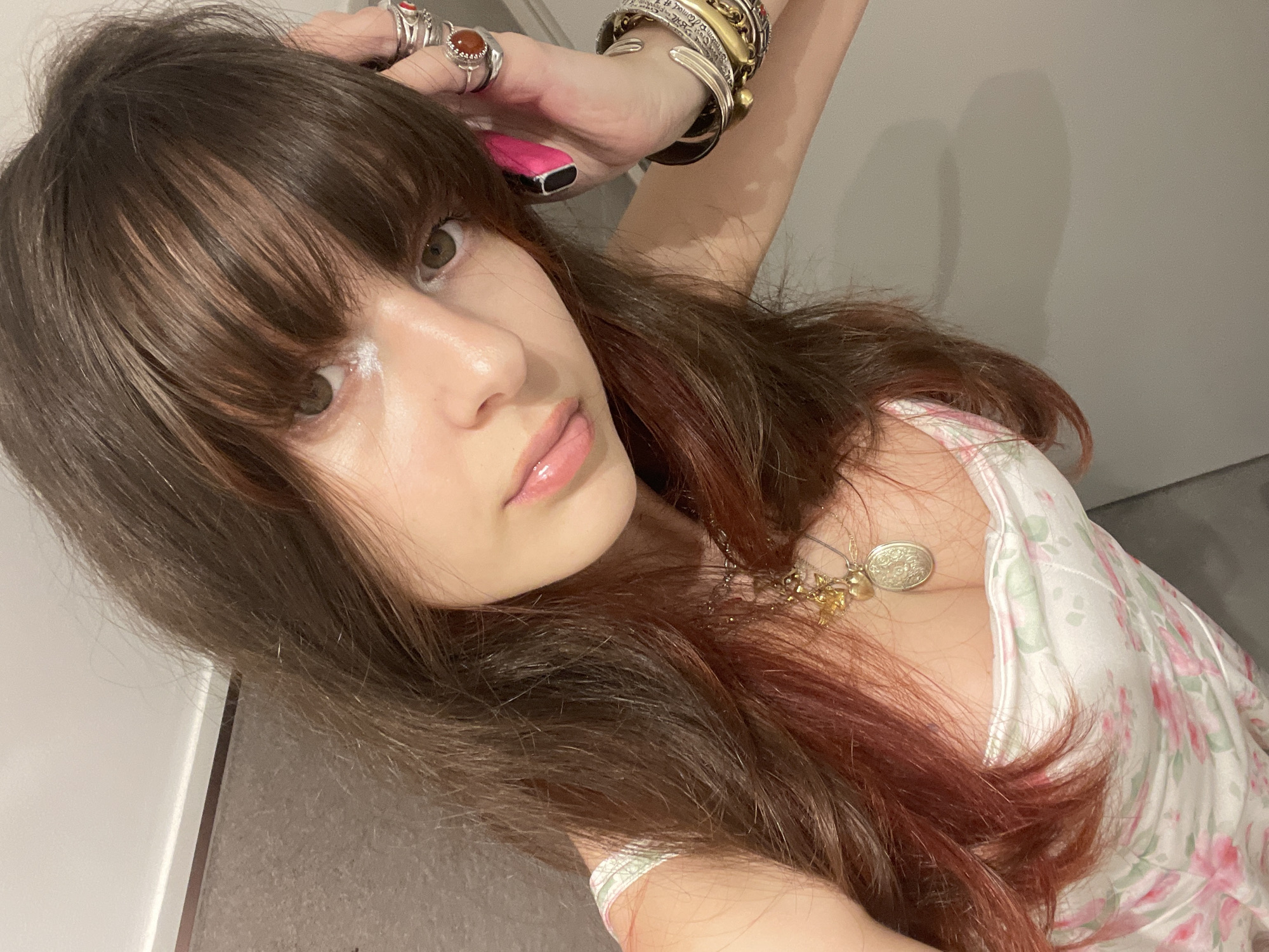 A woman with brown hair and bangs is posing for a selfie. She is wearing a white floral dress and jewelry, including rings, bracelets, and a necklace with a coin pendant. The background appears to be a plain wall, part of which is white and the other grey. There is nothing in the image to place it specifically in Raanana, Israel.