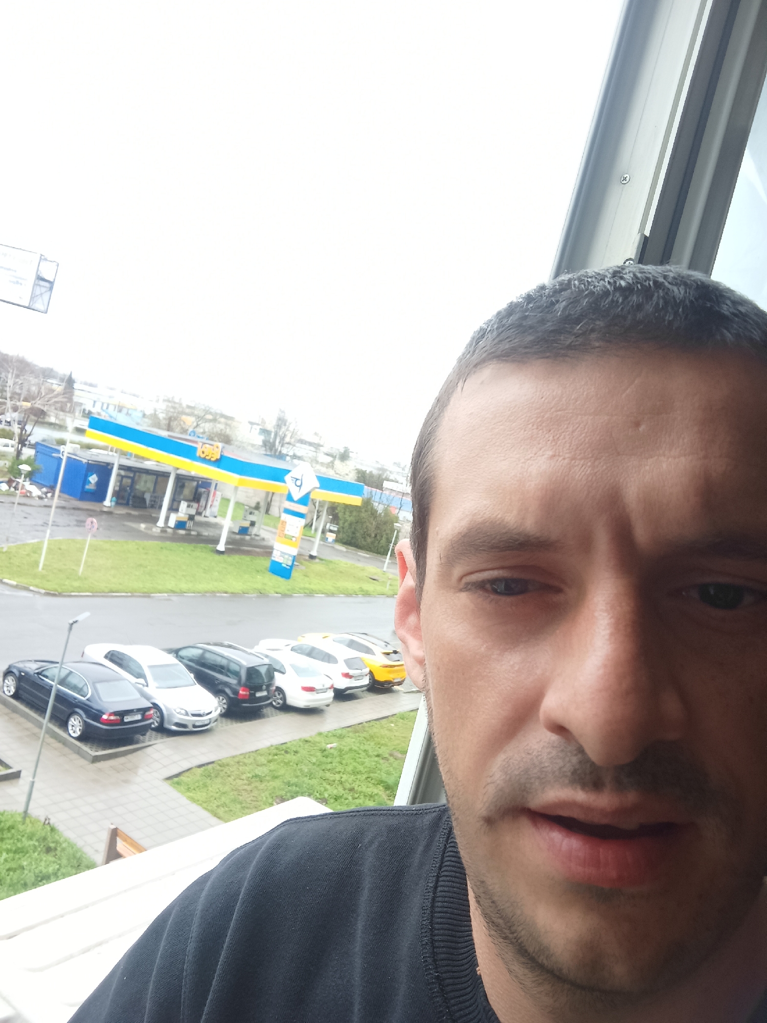 The image is a selfie taken from an elevated position, likely a balcony or high window, looking out over a street scene. In the foreground is a man with short, dark hair, and a dark crewneck shirt. He is positioned to the right of the frame, taking up a significant portion of the image. His expression is somewhat neutral.
Behind him and below, there is a view of a street with several parked cars, including a dark blue sedan, a silver sedan, a dark-colored SUV, a white sedan, and a bright yellow sports car. Across the street is a gas station, identifiable by its blue and yellow canopy. The branding on the gas station has blue and yellow accents which matches the colors of the flag of Ukraine. The gas station is located on a patch of green grass.
The overall environment appears to be an urban or suburban area. The sky is overcast and the light appears soft and diffused. The road surface is wet. Given the architectural style of the gas station and the parked cars, and the Ukrainian colors, it is likely the location is Burgas, Bulgaria.