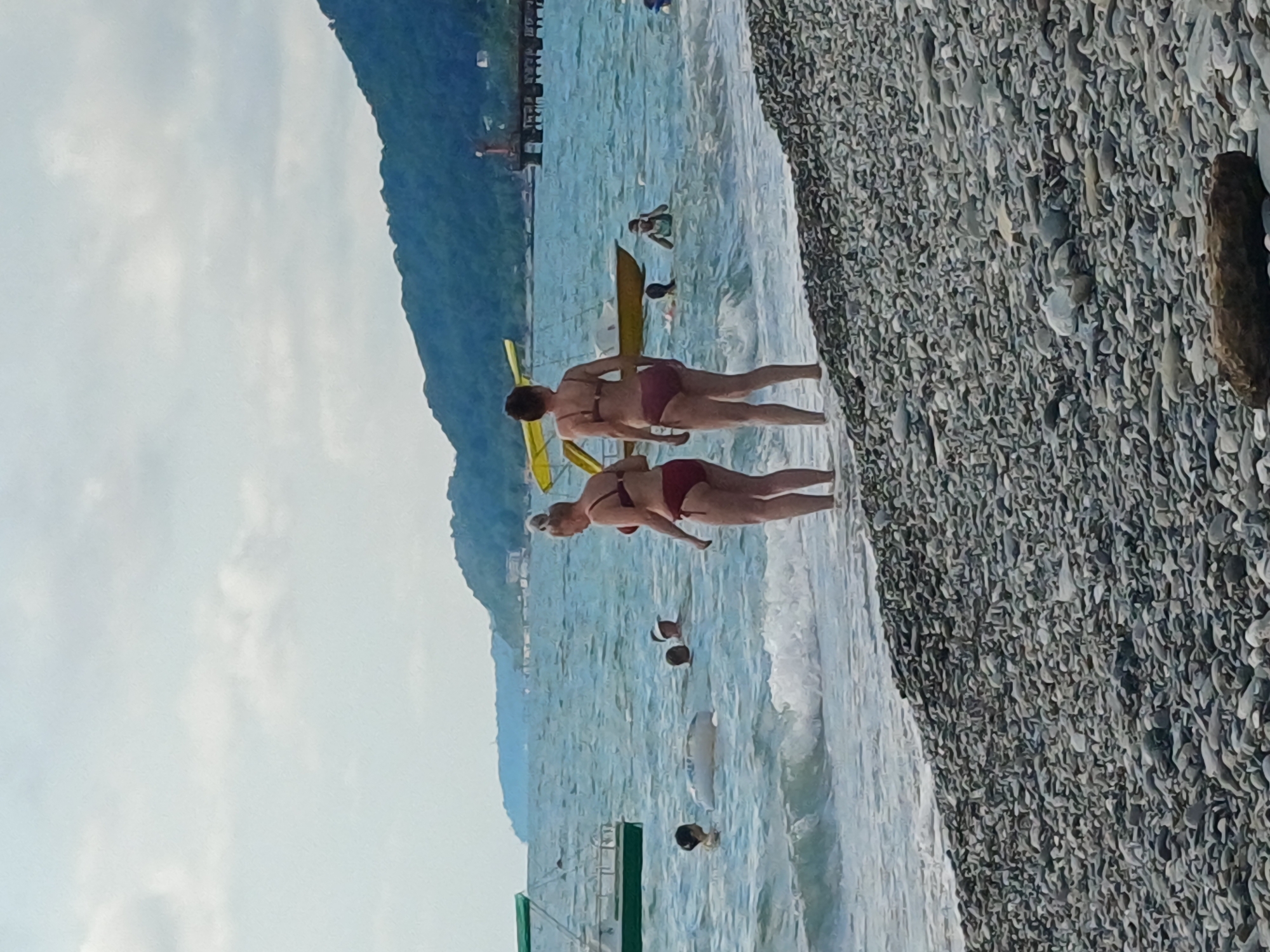 The image depicts a pebble beach in Sochi, Russia, with the sea in the background. The scene is set on a sunny day, as indicated by the bright sky and the clear water. Several people are visible on the beach and in the water. In the foreground, two women in bikinis are standing near the water's edge. One woman is facing away from the camera, while the other is partially visible from the side. Further out in the sea, several individuals are swimming or wading. One person appears to be on a yellow inflatable raft. A dark, rocky mountain range forms the backdrop to the scene, with a pier and some structures visible in the distance on the left side of the image. The overall atmosphere appears to be one of leisure and recreation.