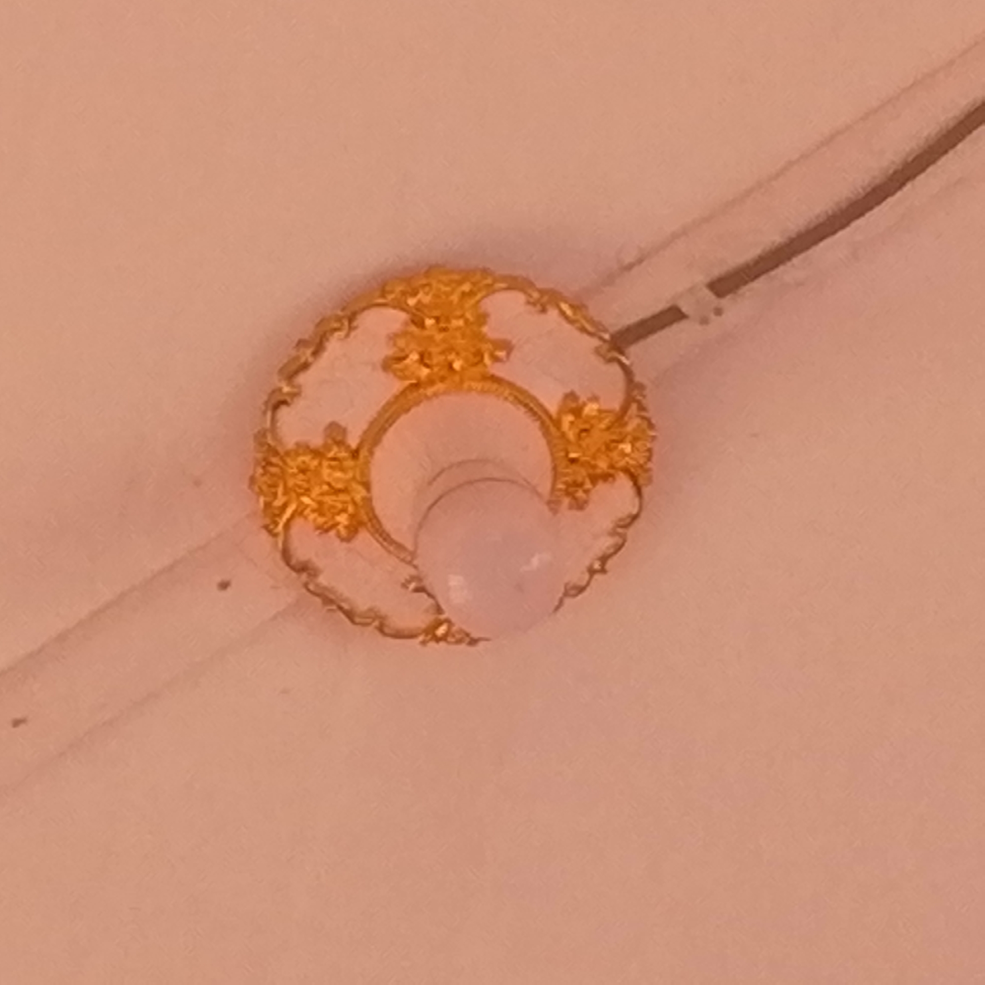 A low-angle shot shows a ceiling light fixture. The fixture has a circular, white base with ornate gold floral details around the perimeter. A rounded light bulb protrudes from the center of the fixture. To the top right and left, a white cable or conduit is visible, appearing to run along the ceiling. The background is a uniform peach color, likely the ceiling itself. The location is Unknown, Unknown.
