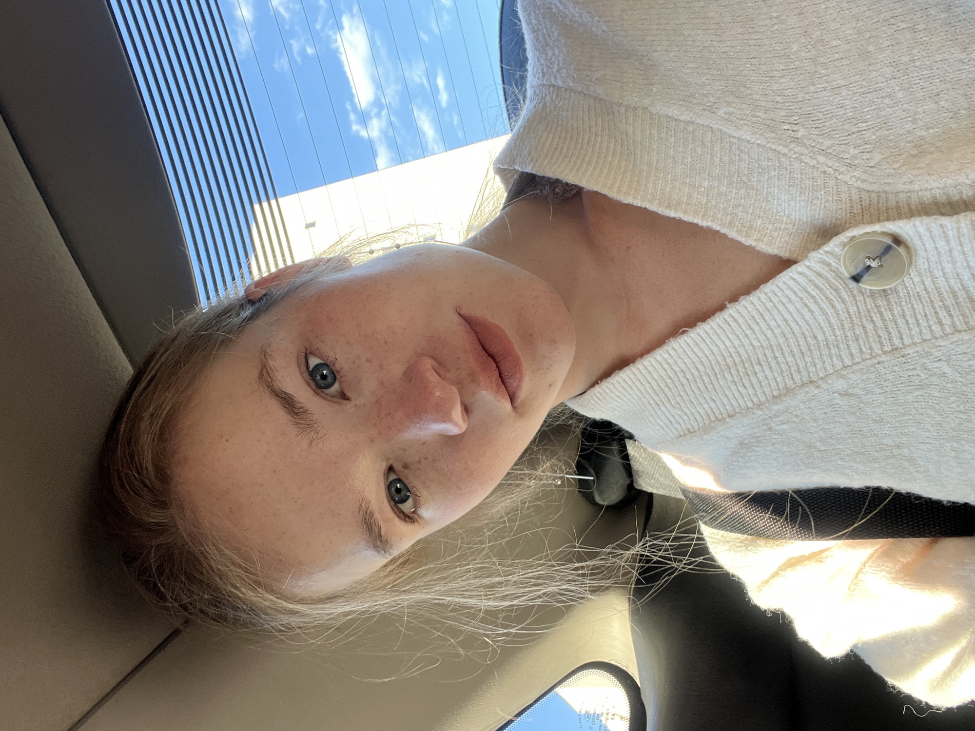 A young woman is photographed from a low angle, likely inside a car. She is fair-skinned with freckles, blue eyes, and light blonde hair. She is wearing a cream-colored sweater with a visible button. The background shows a window with blinds and a partial view of a building and blue sky. The location is unknown, and not possible to know even with reference to the specified town of Boreti, Montenegro. The interior of the vehicle is visible, including the headrest and rearview mirror.