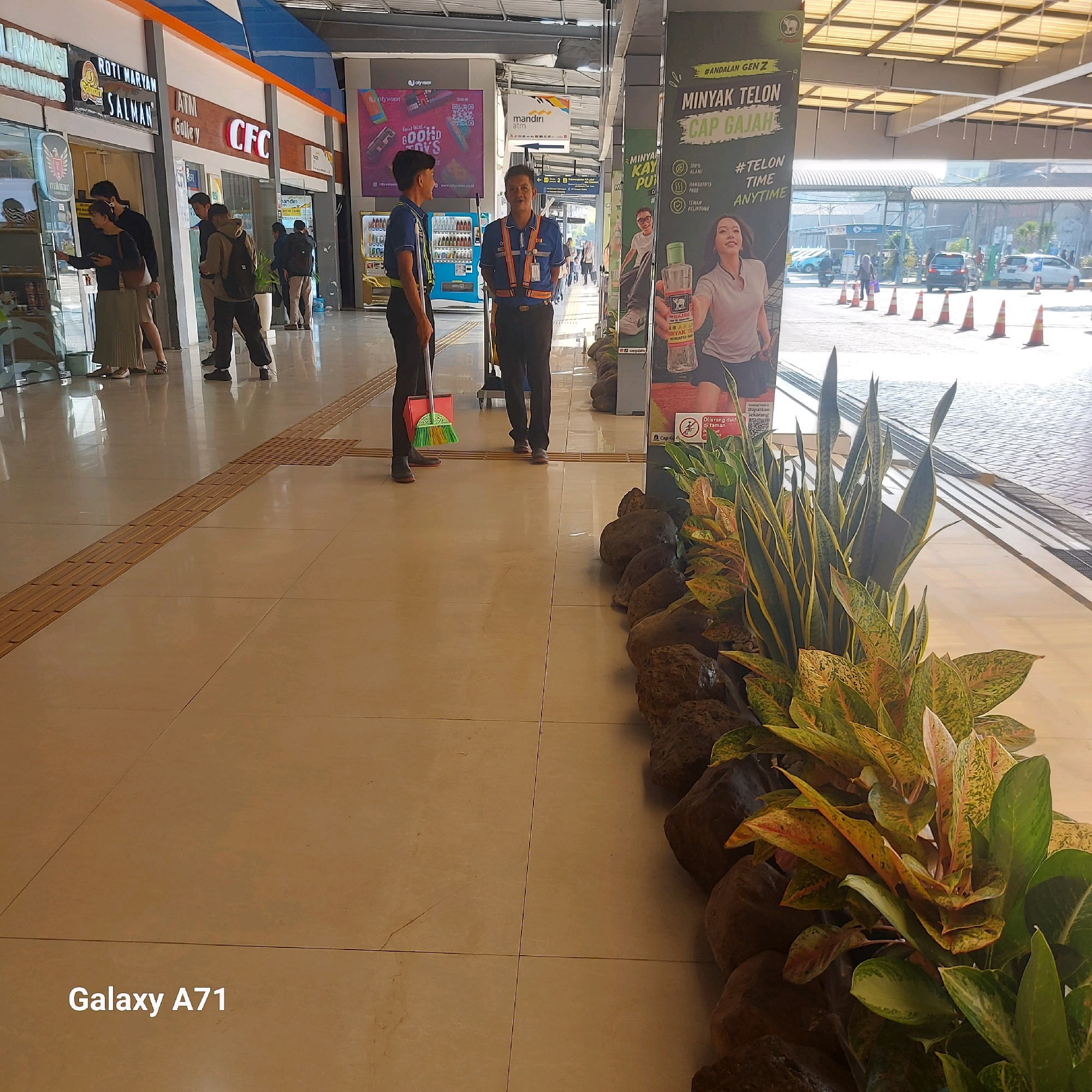 The image depicts a wide, well-lit interior walkway or plaza, possibly within a transit hub or shopping center in Surabaya, Indonesia. The floor is tiled with large, pale beige tiles, with a distinct tactile paving strip running diagonally across the foreground.
On the left side, a row of shops is visible, including one with a sign that reads "ROTI MARYAM SALMAN". A few individuals are seen interacting in front of these shops, dressed in casual attire.
In the center of the walkway, two male figures in uniform are standing. One, in the foreground, wears a blue shirt with orange and white reflective stripes across his chest and shoulders, dark trousers, and a lanyard. He appears to be a security guard or employee. The other figure, slightly to his left and further back, is also in a blue uniform and is holding a broom and a red dustpan, suggesting he is a cleaner.
On the right side, a large advertisement poster is prominently displayed. It features text in Indonesian, including "MINYAK TELON CAP GAJAH" (Elephant Brand Baby Oil), and images of a woman and a product bottle. Next to the poster is a potted plant arrangement with large, broad leaves and a dark stone border.
Further down the walkway, there are glimpses of outdoor areas with parked vehicles and orange traffic cones, suggesting an entrance or exit to a parking lot or road. The overall atmosphere is bright and suggests a public, active space. The "Galaxy A71" watermark in the lower left corner indicates the device used for capturing the image.