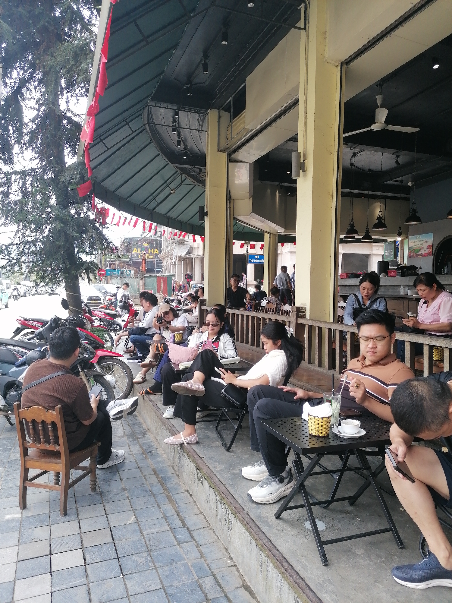 People are sitting outside a building in Sa Pa Ward, Vietnam. There are motorcycles parked in front of them. The building has red and white flags hanging from its roof. Some people are looking at their phones, while others are talking to each other. There is a table with a drink and a cup of coffee on it.