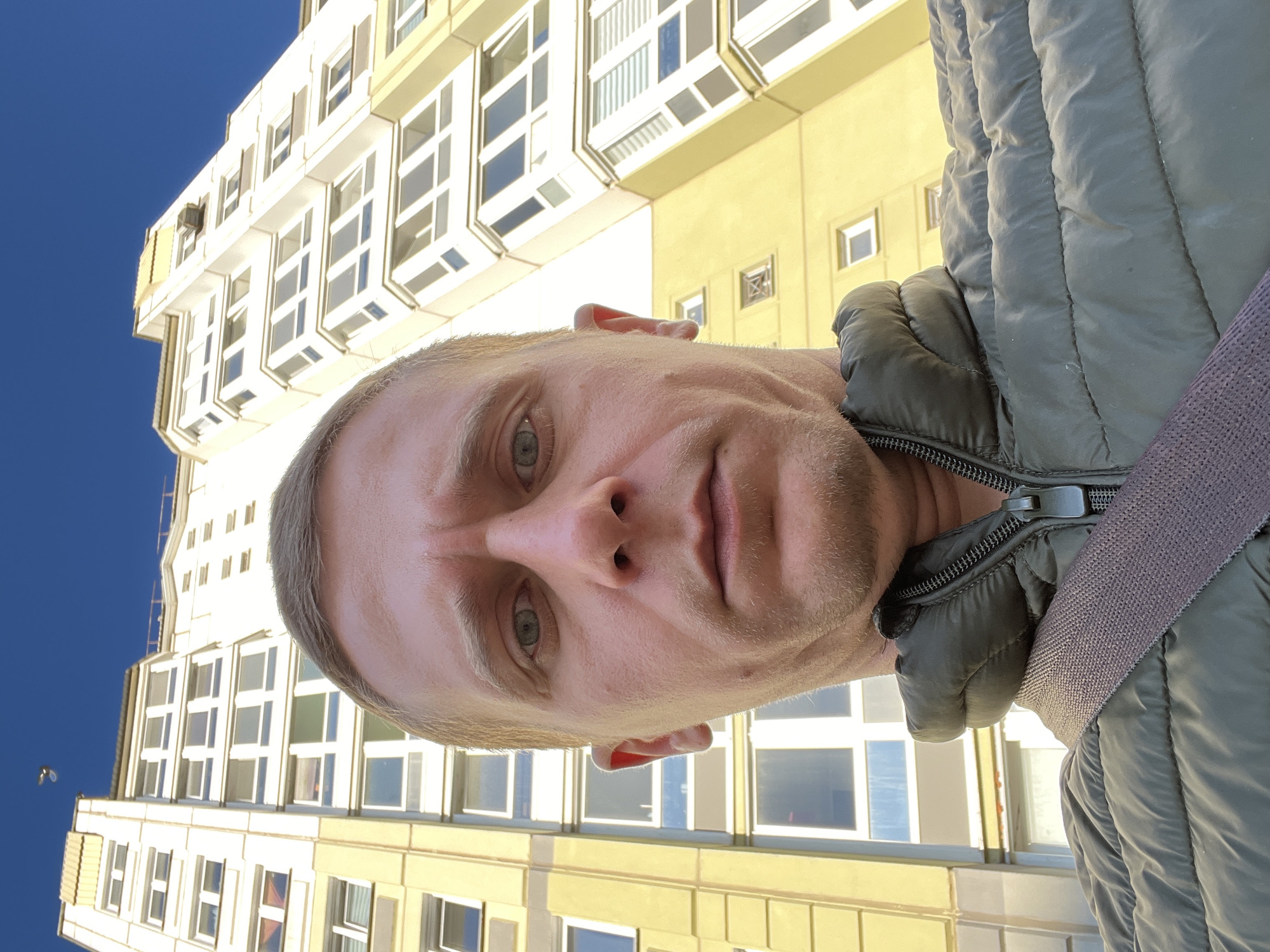 A low-angle, close-up selfie shows a light-skinned man with short, light-brown hair and green eyes. He is wearing a dark green puffer jacket with a grey strap across his shoulder. The man is standing outdoors, with a tall, yellow apartment building in the background. The sky above is a clear, bright blue. In the upper left corner of the image, a small, dark object, possibly a bird, is visible against the sky. The building features numerous windows and balconies. The overall impression is that of a casual snapshot taken on a sunny day in an urban environment.