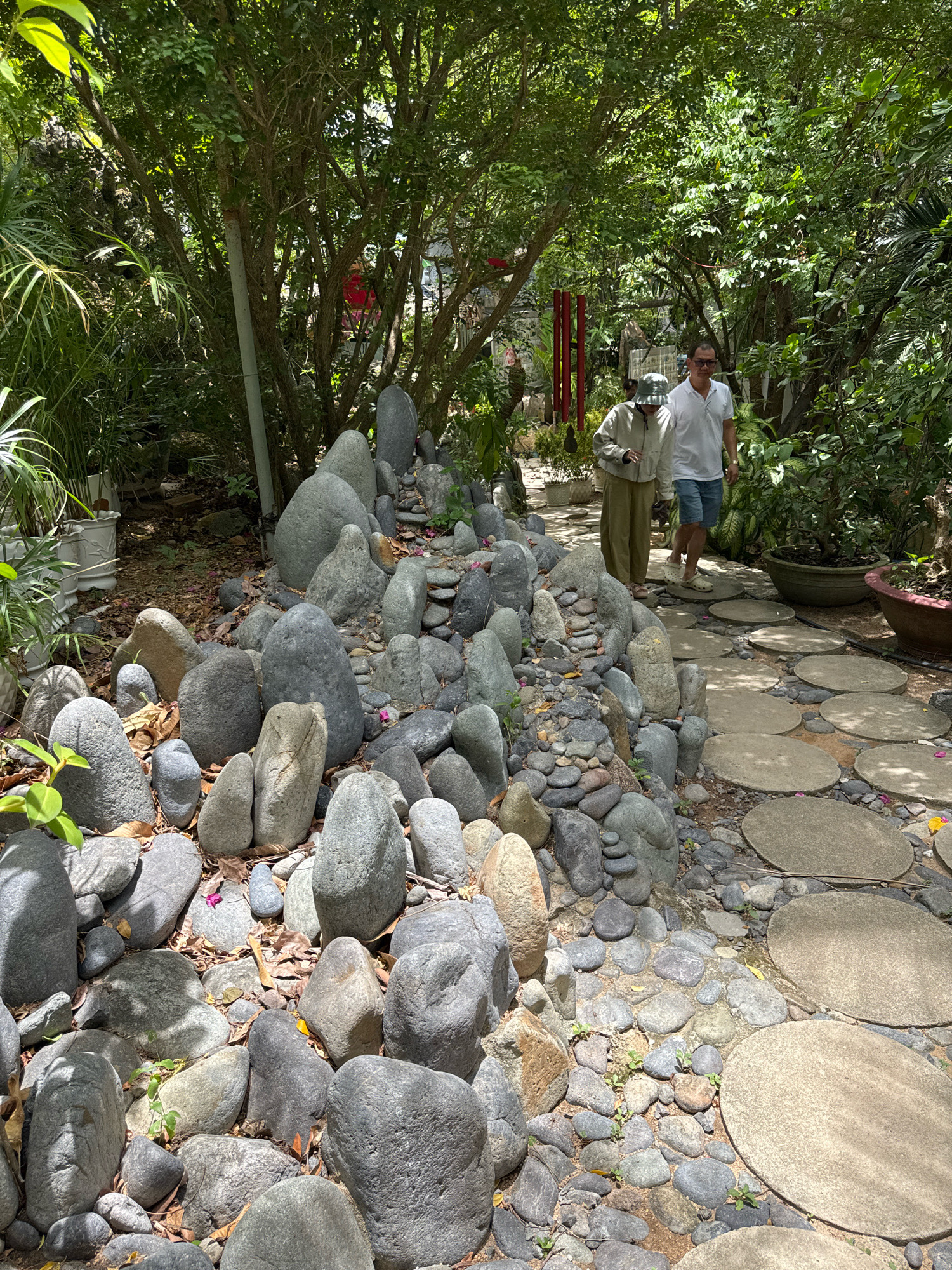 Two people, a man and a woman, walk on a stone path in a garden. The man, wearing a white polo shirt and blue shorts, smiles at the camera. The woman, in a beige outfit and a bucket hat, walks ahead of him. The garden is lush and green, with trees and plants surrounding the path. A pile of grey rocks is to the left of the path. The scene is located at a Buddhist temple in Nha Trang, Vietnam.
