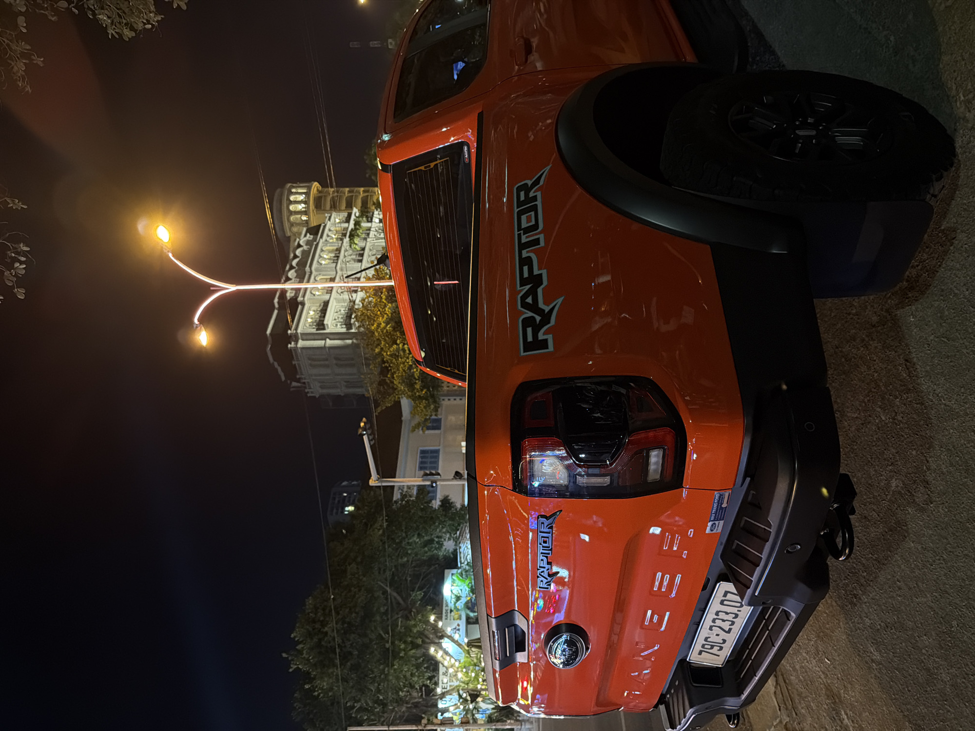 The image shows the rear passenger side of an orange Ford Ranger Raptor pickup truck parked on a street at night in Nha Trang, Vietnam. The truck has a black fender flare and a black mud flap. The license plate reads "79C-233.07". In the background, there are trees, buildings, and a street lamp. One of the buildings appears to be a hotel with ornate architecture. The street is paved and appears to be a sidewalk or curb area. The overall scene depicts a typical urban streetscape at night, with the truck being the primary subject.