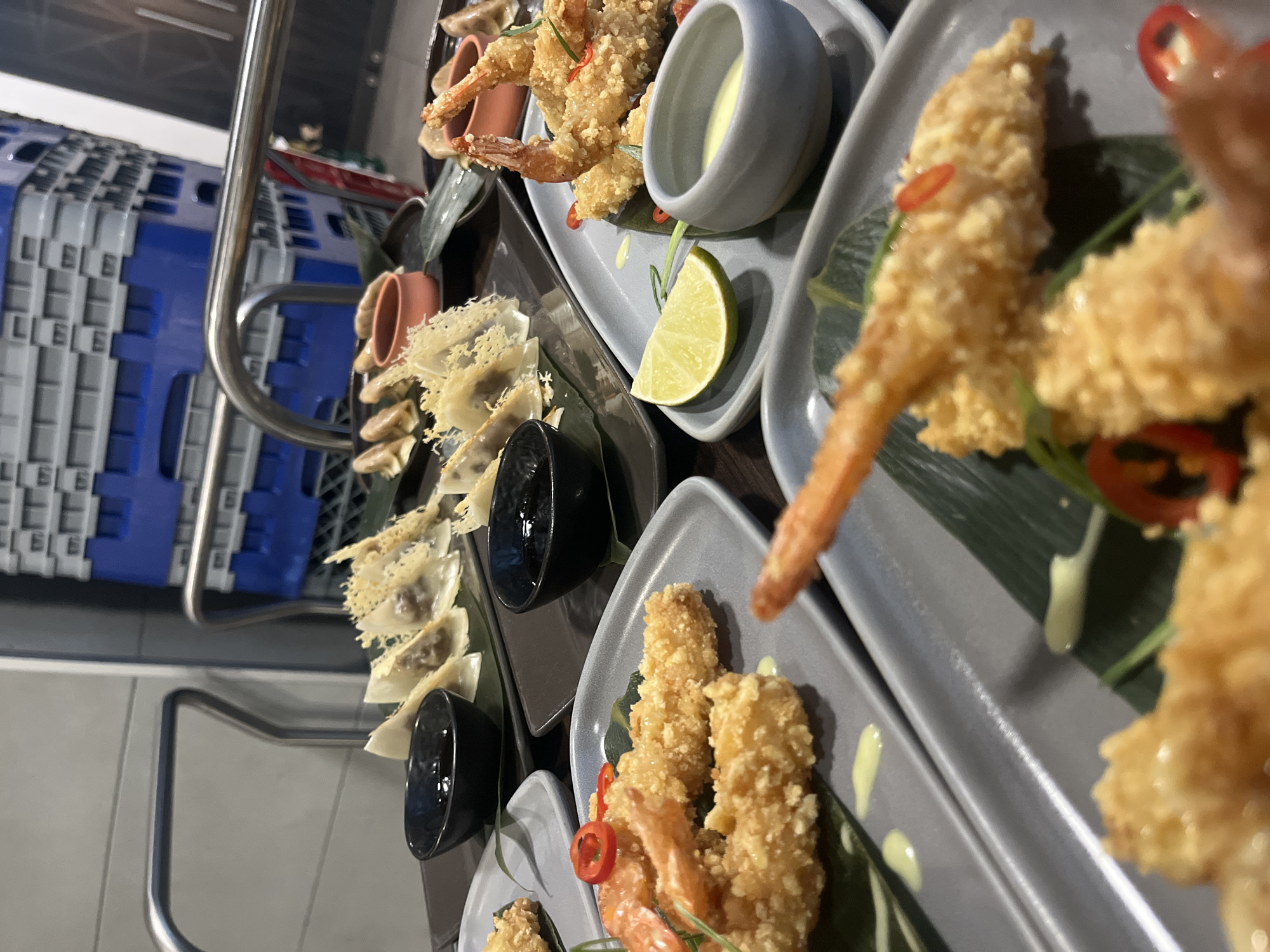The image shows multiple plates of Asian-inspired cuisine, possibly tempura shrimp and dumplings, arranged on a metallic cart. The background includes industrial elements such as the cart itself and what appears to be a stack of blue and grey crates or containers. The food is styled with garnishes like lime wedges and chili peppers. The location information (Sofia, Bulgaria) is not evident from the visual content of the image alone.