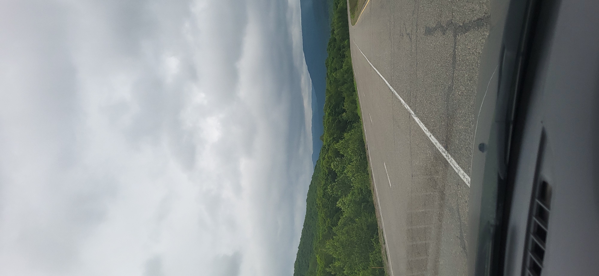 The image shows a road in Zanadvorovka, Russia, with lush green hills and mountains in the background. The sky is overcast with dark, heavy clouds, suggesting a stormy or rainy day. The road is asphalt and has white lines marking the lanes. The foreground shows the dashboard of a car, indicating the photo was taken from a moving vehicle. No people or other subjects are visible in the image. The environment is natural and appears to be a rural or mountainous area.