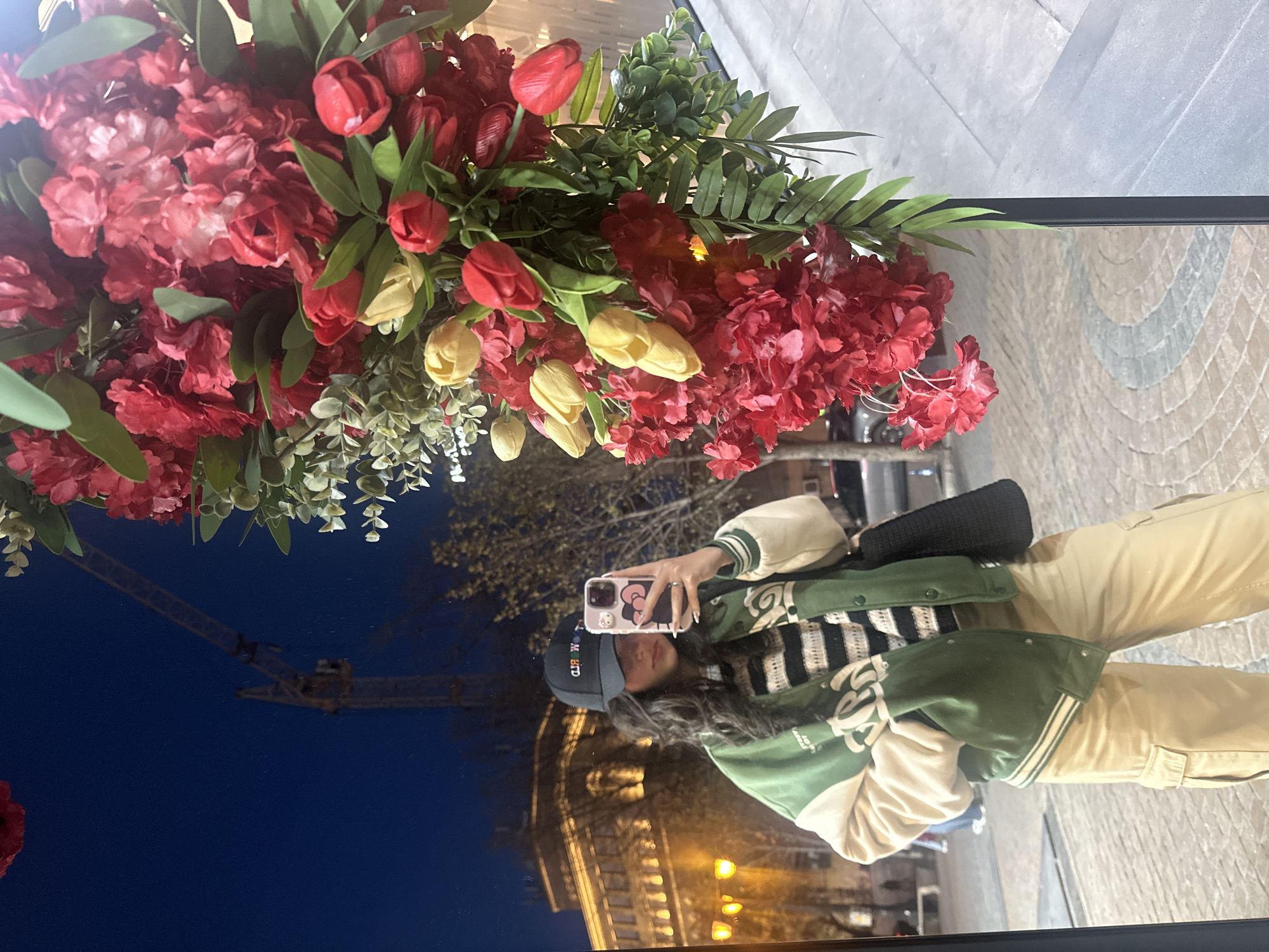 A young woman is taking a selfie in a mirror display decorated with red and yellow flowers. She is wearing a baseball cap, a green and white varsity jacket, and khaki pants. The mirror reflects a street scene at night, featuring a building with architectural lighting and a construction crane. The location appears to be Yerevan, Armenia.