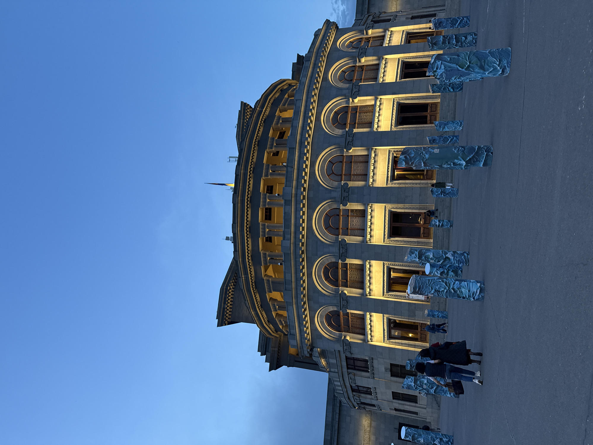 Here is a description of the image:
The image shows the National Gallery of Armenia in Yerevan at dusk. The building's curved facade is illuminated, contrasting with the darkening blue sky. Below, the plaza features a row of sculptures, and several people are visible. The composition is oriented vertically.