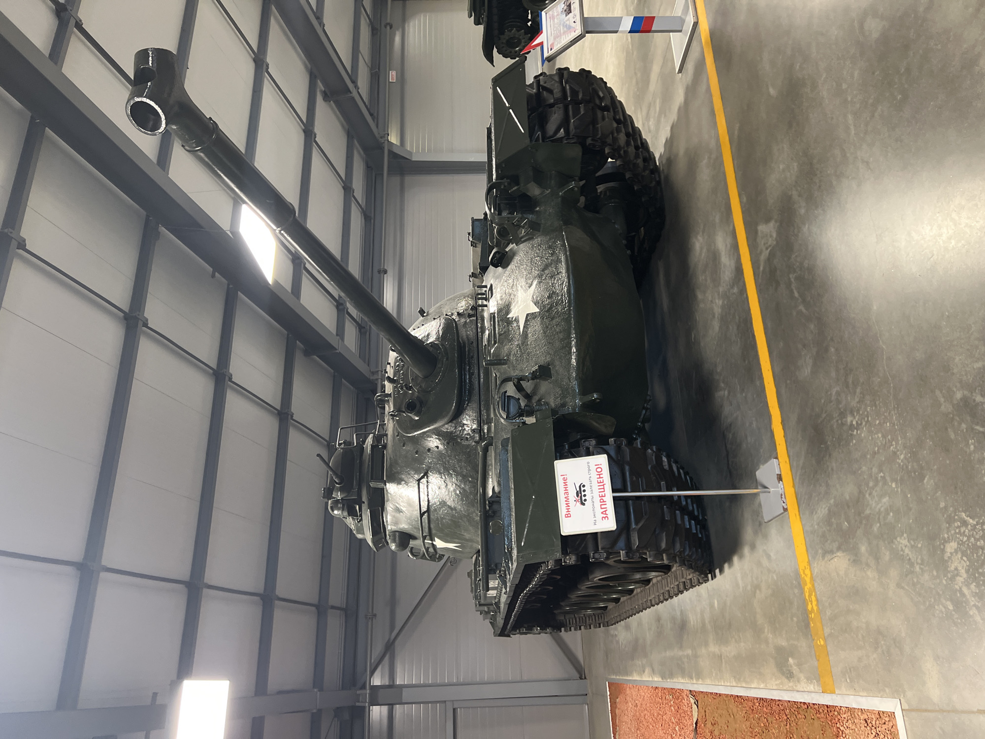 A dark green, American M47 Patton tank is displayed indoors at the Patriot Exhibition Center in Kubinka, Russia. The tank is positioned centrally in the frame, facing slightly to the left. Its long cannon barrel extends diagonally upwards, nearly touching the ceiling structure. A white sign with red text, reading "Внимание! На экспонаты залезать строго ЗАПРЕЩЕНО!" (Attention! Climbing on the exhibits is strictly FORBIDDEN!), is placed in front of the tank's left track. The tank features white star insignia on its hull. The environment appears to be a large, well-lit exhibition hall with a corrugated metal ceiling supported by dark grey beams. The floor is concrete, with a yellow line painted on it running horizontally across the foreground. To the left of the tank, a section of ground covered in reddish-brown gravel is visible, separated by a metal barrier. A small, informational stand with Russian flag colors is positioned to the right of the tank's front. No people are visible in the image.