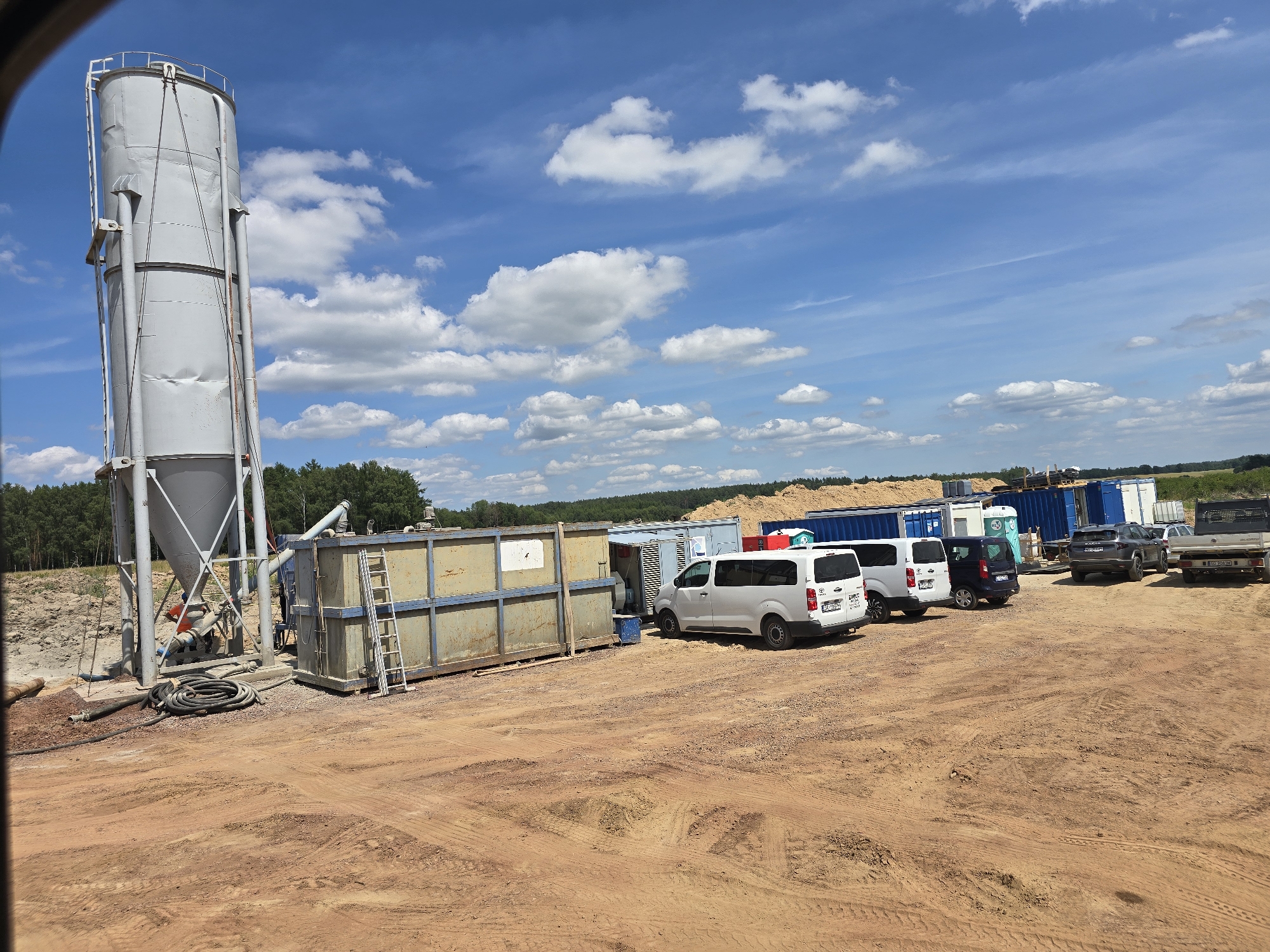 This image depicts an outdoor scene at what appears to be a construction or industrial site in Piecnik, Poland. The central focus is a large, grey cylindrical silo, likely for storing materials, which is situated on the left side of the frame. Adjacent to the silo is a long, metallic rectangular container with a ladder attached, suggesting it is used for storage or as part of a processing unit. Several vehicles are parked in the background, including white vans, a dark-colored SUV, and a pickup truck. Interspersed among the vehicles are shipping containers, some blue and some white. Piles of material, possibly wood or sand, are visible further back, along with a line of trees forming the distant horizon. The ground is composed of reddish-brown dirt with tire tracks, indicating recent vehicle activity.
The sky is bright blue with scattered white cumulus clouds, suggesting fair weather and daytime, likely mid-morning to afternoon. The sunlight casts shadows, further indicating the time of day. There is no clear activity or event taking place; the scene appears to be a static view of the site.
Notable details include the weathered appearance of the silo and the storage container, hinting at the functional nature of the location. The presence of various containers and vehicles suggests logistical operations. Visible text is limited; a license plate is partially visible on one of the vans, and a logo or small label is on the back of another van. No people are clearly visible in the image. The overall impression is one of an active, albeit currently quiet, industrial or construction yard under a pleasant sky.