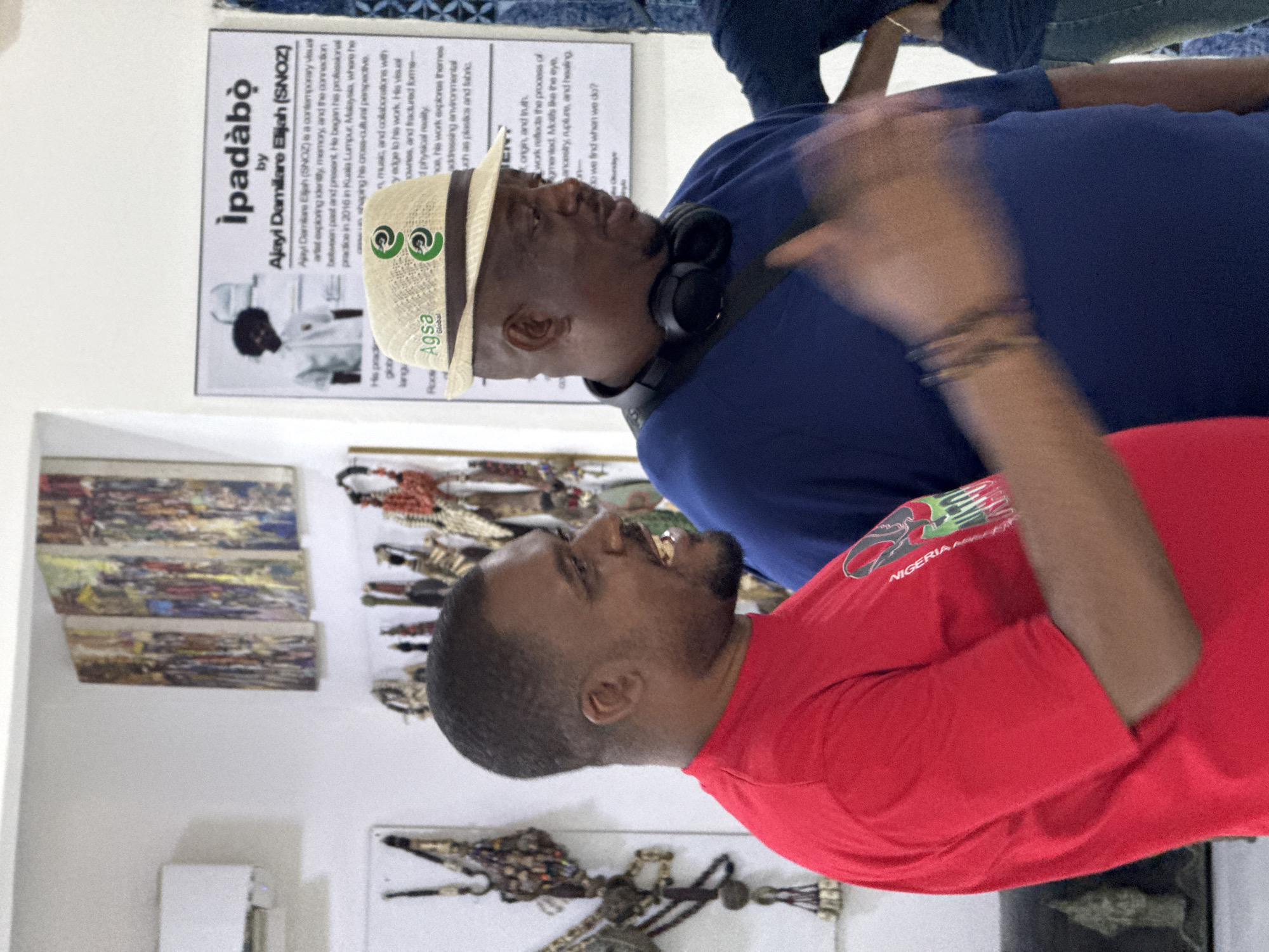 This image captures two men engaged in conversation at what appears to be an art exhibition or gallery, likely in Abuja, Nigeria. The scene is brightly lit indoors, with white walls displaying various artworks.
In the upper part of the frame, a dark-skinned man wears a blue short-sleeved shirt, a straw fedora-style hat with a green "Agsa Global" logo, and black over-ear headphones around his neck. He looks downwards, seemingly at the second man, with his right arm raised and blurred, indicating movement or a gesture during discussion.
Facing him, in the lower part of the frame, is another dark-skinned man wearing a red short-sleeved t-shirt. Visible on his shirt is part of a green and white logo that includes the word "NIGERIA". He looks upwards with an animated expression, mouth slightly open, suggesting he is actively speaking or reacting to the other man.
On the wall behind them, a prominent poster is titled "Ipadabò by Ajayi Damilare Elijah (SNOZ)". The text identifies Ajayi Damilare Elijah (SNOZ) as a contemporary visual artist who explores themes of identity, memory, the connection between past and present, and a cross-cultural perspective, with professional practice beginning in Kuala Lumpur, Malaysia. His work addresses environmental concerns like plastics and fabric, and delves into ancestry, rupture, and healing. Other artworks, including three horizontal pieces and a sculptural piece, are visible on the walls.
The interaction between the two men, set against the backdrop of the exhibition poster and artworks, suggests a focused discussion about the art or the artist's themes, possibly during an exhibition viewing or artist talk.