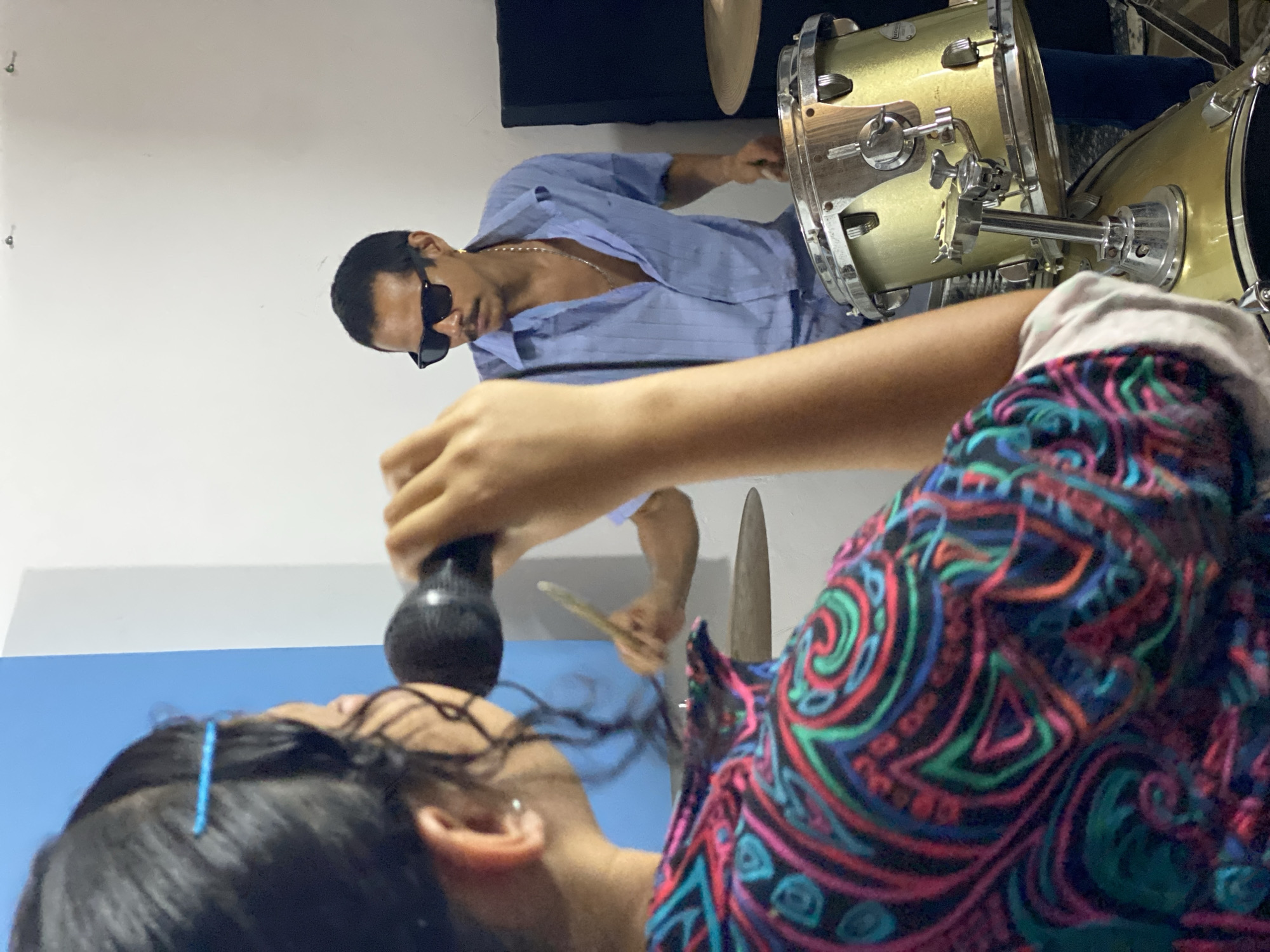 The scene appears to be indoors in Tinaquillo, Venezuela. A man wearing sunglasses and a light blue open shirt is playing a gold-colored drum kit. In the foreground, another person holds a microphone near their mouth; they are wearing a colorful patterned shirt.