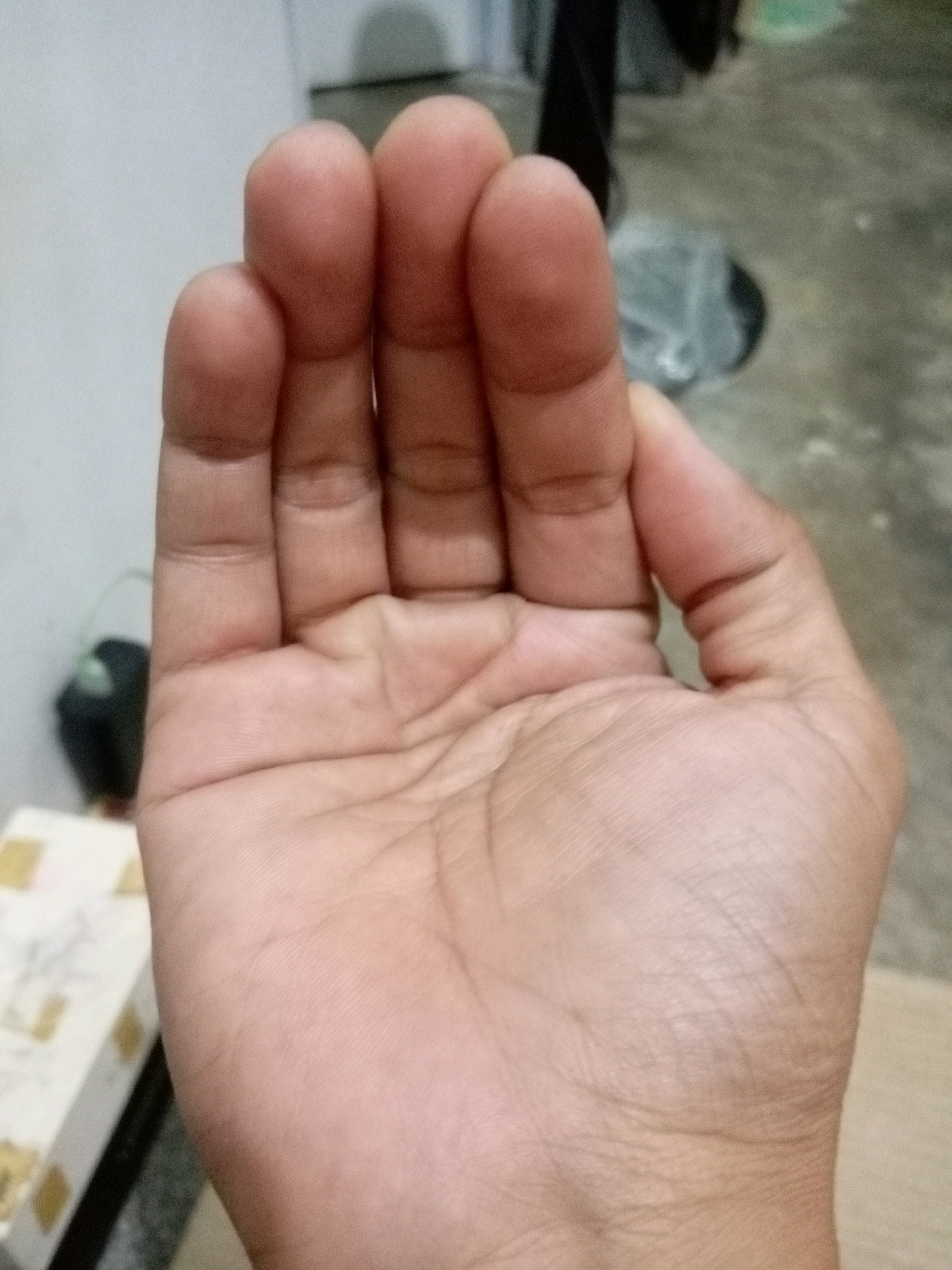 A close-up, eye-level shot displays the palm of a person's hand with their fingers partially curled inwards, obscuring the palm. The fingers, from left to right, appear to be the thumb, index finger, middle finger, and ring finger. The skin tone is light to medium. The background is out of focus but suggests an indoor environment with a concrete floor, some stacked boxes with a patterned design on the left, and a dark object in the middle ground. The scene is devoid of any context that would place it in Batam, Indonesia, or any other specific city.