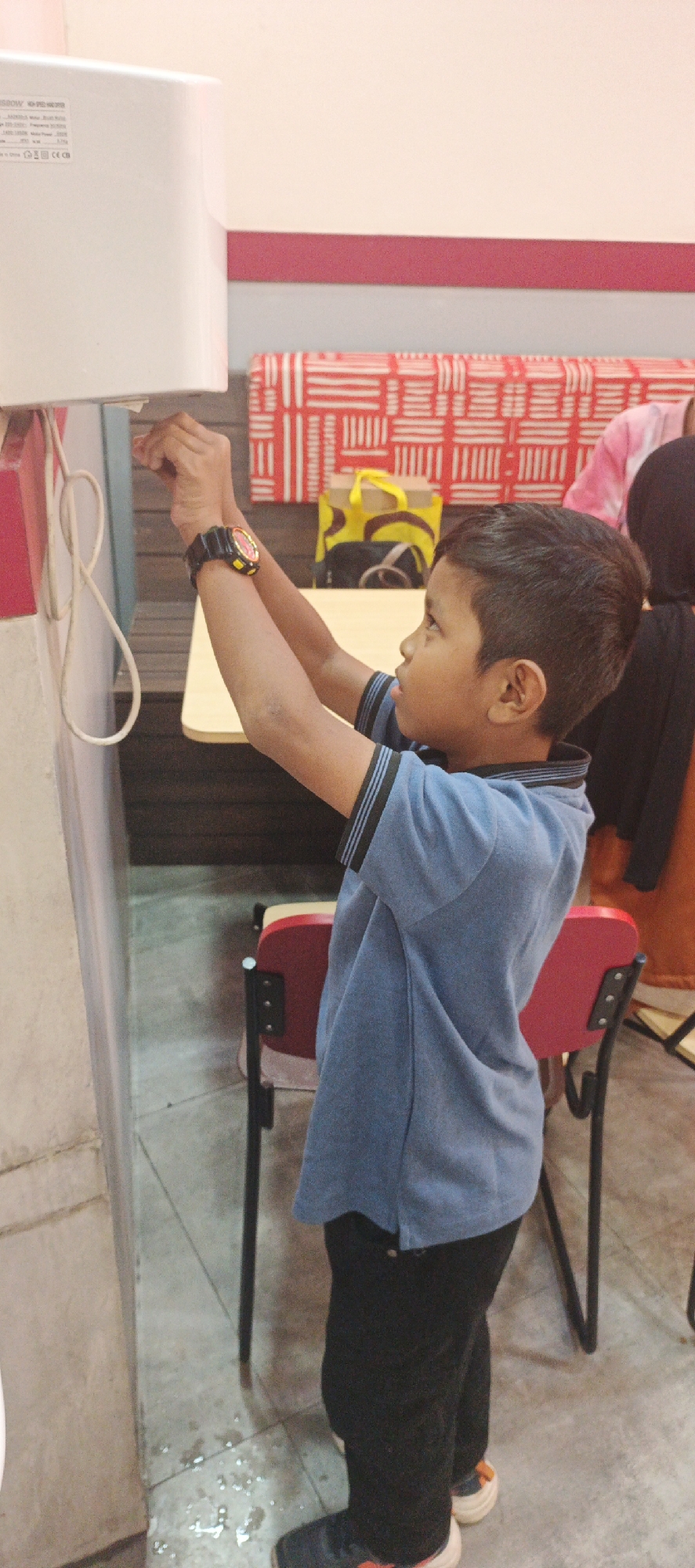 A young boy is depicted in a medium shot, standing indoors and reaching up to interact with a white automatic hand dryer mounted on a wall. He is wearing a blue polo shirt with dark stripes on the collar and sleeves, and black pants. On his left wrist, he wears a black digital watch with a red and yellow face. His hands are positioned near the sensor or activation mechanism of the hand dryer.
The background reveals a room with a wooden table and red chairs. A patterned red and white fabric is visible on a wall or partition behind the table. A yellow shopping bag is placed on the table. The floor appears to be tiled and slightly wet, with small puddles of water. The overall lighting suggests an indoor setting, possibly a public restroom or a facility with hand-washing stations.
While the image provides details of the boy's actions and the immediate environment, it does not contain any explicit information that would allow for the determination of its specific location within the city of Surabaya, Indonesia.