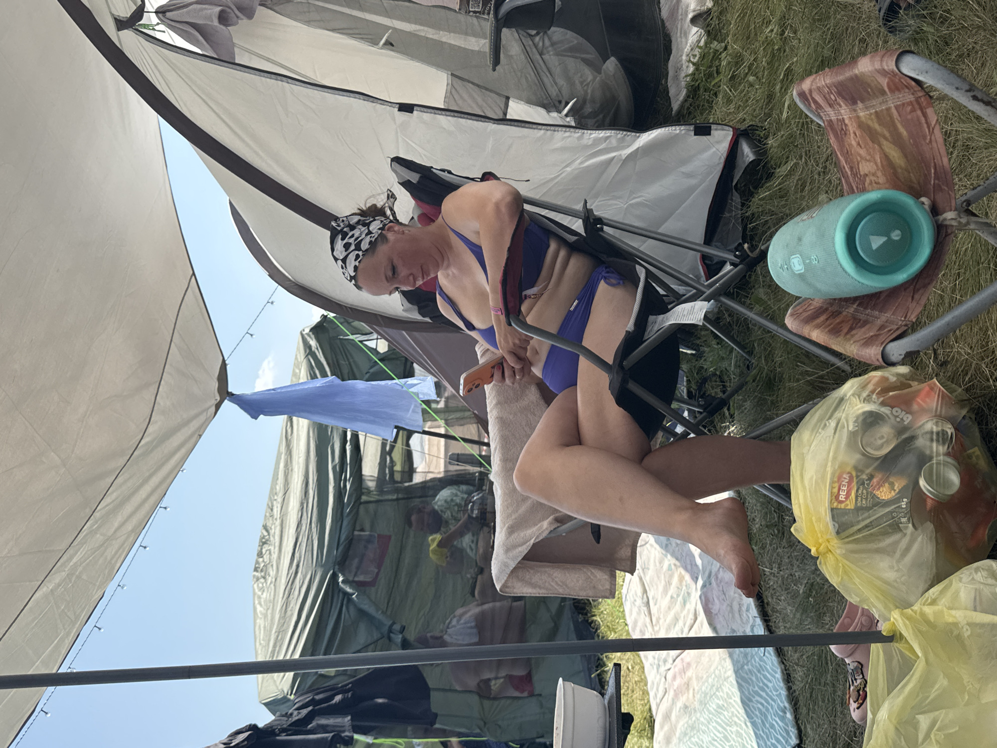 A woman sits in a camping chair under a canopy, looking at her phone. She is wearing a purple bikini and a bandana on her head. She is in a grassy area with tents and camping equipment around her. In the background, two men are visible inside a tent. There are also yellow trash bags filled with cans. A blue portable speaker rests on another camping chair. The weather is sunny.
