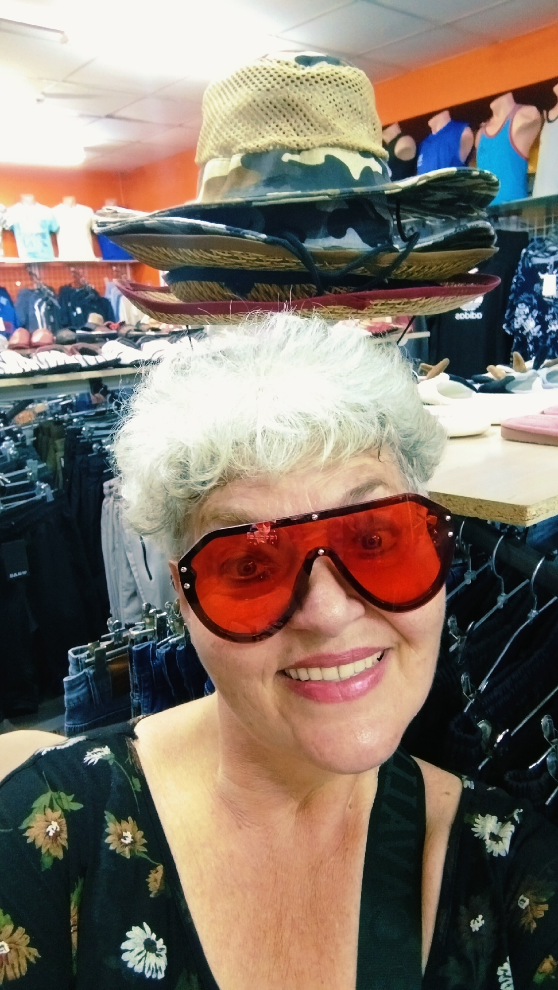 A woman with short, curly, grey hair and red-tinted aviator sunglasses smiles at the camera. She is wearing a dark floral print top. The background appears to be a retail store, with clothing racks visible. On the top of her head, she has stacked several hats. The scene is indoors with artificial lighting.