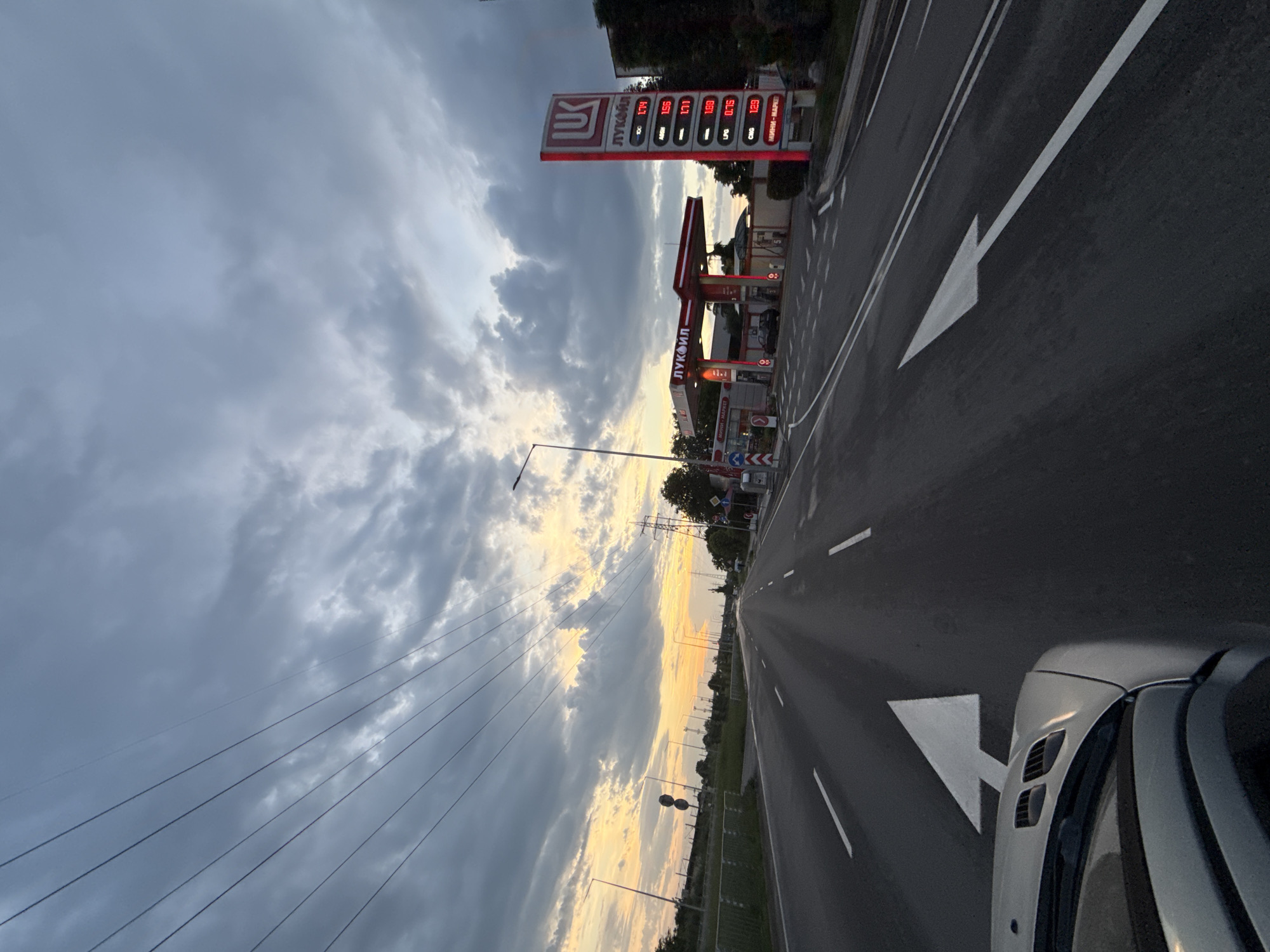 The image captures a view from a vehicle on a road, likely within a city, evidenced by the presence of a Lukoil gas station sign. The sign displays fuel prices, including those for "100," "Дизел," "Бензин-95," "Бензин-100," "LPG," and "CNG," among others, with the term "МИНИ-МАРКЕТ" also visible. The road has directional markings, including a white arrow pointing left. Overhead, a series of power lines stretch across the frame, contrasting against a cloudy sky illuminated by the setting sun. The car's exterior is partially visible in the lower right corner of the image. Based on the presence of the Lukoil gas station sign, which is common in Eastern Europe, and specifically the use of Cyrillic script on the sign, it's plausible the location is within Bulgaria, but further identifiers such as the local currency on the price display or unique architectural styles would be required to definitively determine the exact city.