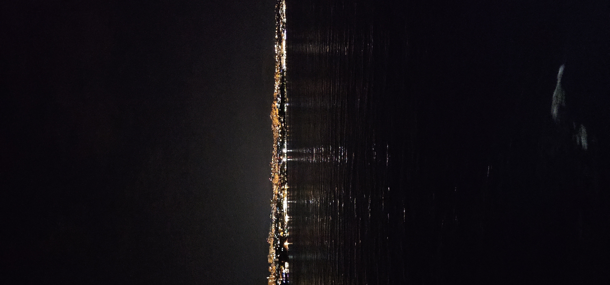 The image depicts a nighttime view of the coastline of Split, Croatia. A city skyline, illuminated by numerous lights, stretches across the horizon. The lights are a mix of warm yellow and white, indicating buildings and streetlights. The city is situated on a landmass that meets the dark, rippling water in the foreground. The water reflects the city lights, creating shimmering streaks across its surface. The sky above is dark and largely featureless, suggesting it is either night or heavily overcast. The lower portion of the image is dominated by the dark water, with a faint, pale form visible near the bottom right, possibly a reflection or a submerged object. No people or specific activities are discernible. The scene conveys a sense of urban tranquility at night, viewed from across the water.
