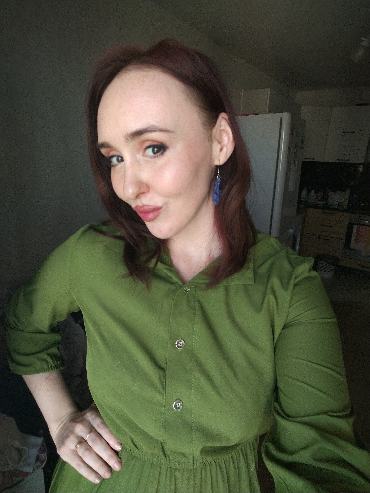 A woman with fair skin, freckles, and auburn hair is posing indoors. She is wearing a long-sleeved, olive-green dress with buttons down the front. She has an earring on her left ear. Her hair is parted to the side and falls to her shoulders. Her left hand is on her hip, and she is pouting her lips slightly. In the background, there is a refrigerator and kitchen cabinets, indicating a domestic setting. There is no information to indicate this image was taken in Izhevsk, Russia.