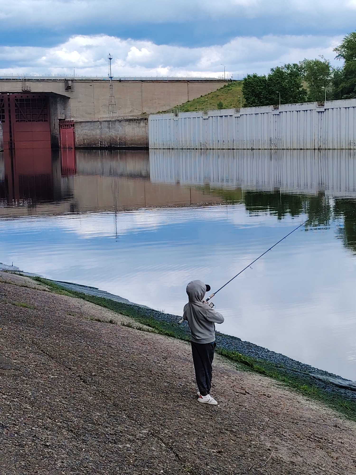 A person is fishing in a body of water near a large concrete structure under a cloudy sky in Chaikovsky, Russia.
The person, likely a young individual, is wearing a gray hooded sweatshirt, dark pants, and white sneakers. They are holding a fishing rod and appear to be in the act of casting or reeling in their line. The fishing rod extends out over the water.
The body of water reflects the cloudy sky and the concrete structure behind it. The structure is a large dam or lock system with red gates and white corrugated walls. It has a tower extending up from its roof. On the far right are some green trees.
The ground where the person is standing appears to be a concrete bank. A small amount of green foliage is growing at the water's edge. The overall lighting suggests an overcast day.