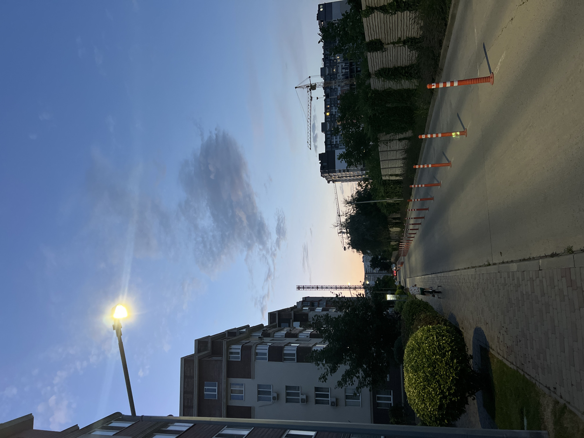 An exterior, eye-level, long shot captures a quiet street scene in Kryzhanivka, Ukraine, during twilight. A tall, modern apartment building with a brick-patterned upper section and white walls dominates the left side of the frame. The building features numerous windows, some with air conditioning units. A brightly lit street lamp extends from the building, casting a warm glow against the darkening blue sky with scattered clouds.
To the right of the apartment building, a paved walkway is bordered by a manicured, rounded shrub and grass. Further along the walkway, a child is riding a bicycle. The street itself is lined with orange and white traffic delineating poles, casting long shadows on the asphalt. In the background, construction cranes are visible above a multi-story building, suggesting ongoing development. Beyond that, more apartment buildings and trees are visible under the twilight sky. The overall impression is one of a developing urban residential area at the end of the day.