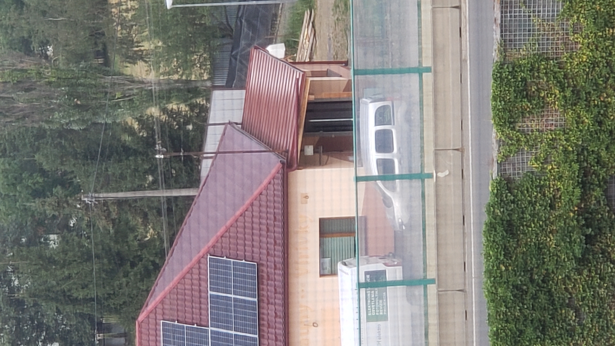 The image captures a residential building with a red-tiled roof adorned with solar panels. A small annex with a similar red roof extends from the main structure. In front of the building, partially obscured by a green fence and concrete barriers, are two vehicles: a white van with lettering that appears to be "IH elektro" and a silver minivan. The background features a dense backdrop of trees and vegetation, suggesting a semi-rural or suburban setting. The location is identified as Prešov, Slovakia.