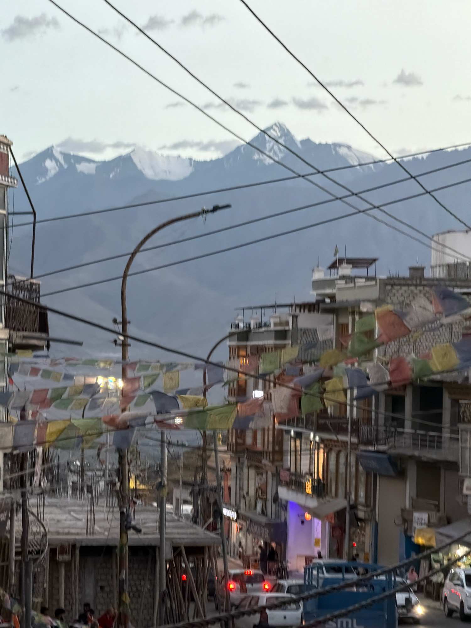 This is an eye-level, medium shot of a street scene in the city of Leh, India, with snow-capped mountains in the background. The sky is overcast with a few clouds. The foreground is dominated by a street lined with multi-story buildings, many of which are adorned with colorful prayer flags. The buildings have a mix of architectural styles, with balconies, windows, and some showing signs of ongoing construction. Several vehicles, including cars and a blue truck, are visible on the street, suggesting moderate traffic. A lamppost with visible wiring is present in the mid-ground. A few people are visible on the sidewalks and near the buildings, but their actions are indistinct. The overall impression is that of a bustling urban environment set against a majestic natural backdrop.