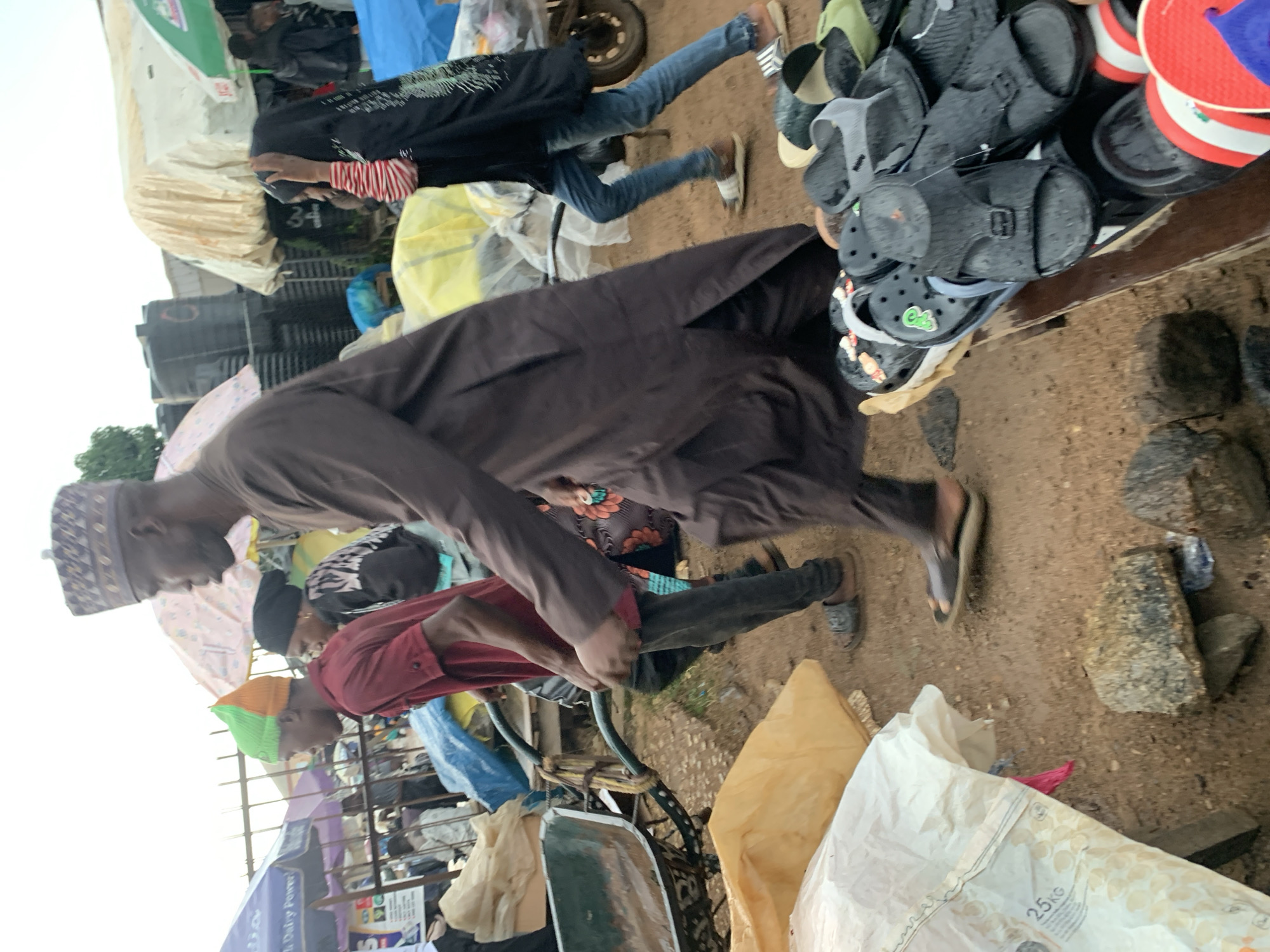In Gura Top, Nigeria, a market scene unfolds with a group of individuals navigating a bustling environment. A man in a brown robe and patterned cap walks towards the right, his gaze directed forward. To his left, a young person wearing a red shirt and an orange and green beanie pushes a wheelbarrow. Behind them, a person in a black headscarf and patterned jacket, and another in a black headscarf and a black outer garment with a red and white striped sleeve, are also present. The ground is sandy and uneven, scattered with rocks and various items, including a large white bag with "25KG" printed on it. In the foreground, a wooden table displays an assortment of footwear, primarily sandals and flip-flops, some appearing wet. Several stalls with umbrellas and tarpaulins are visible in the background, suggesting a vibrant marketplace.