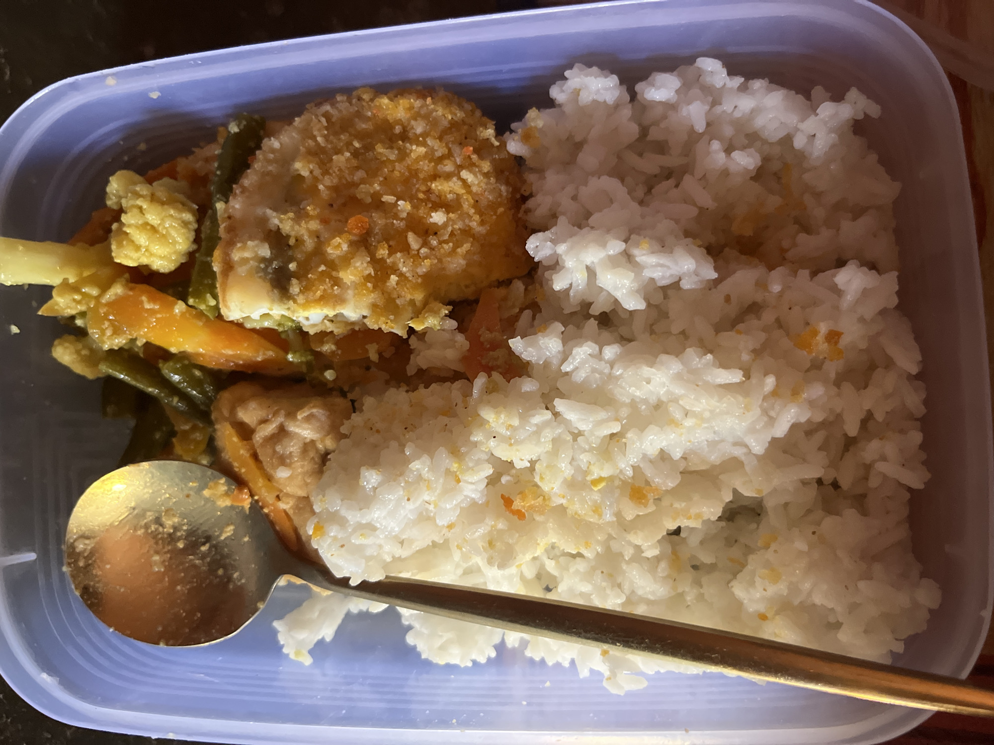 A close-up, top-down shot shows a meal in a light blue plastic container. The meal consists of a generous portion of white rice, with small orange specks scattered throughout. A golden spoon rests on top of the rice, angled from the top left towards the bottom center. To the right of the rice, there is a dish with breaded food, a piece of which has been broken off. Behind this, a medley of vegetables, including green beans, carrots, and cauliflower, are visible, mixed with some chicken or tofu. The lighting suggests it may be indoors. This image does not contain any recognizable landmarks or features that would allow for a precise location within Kruengmate, Indonesia to be identified.