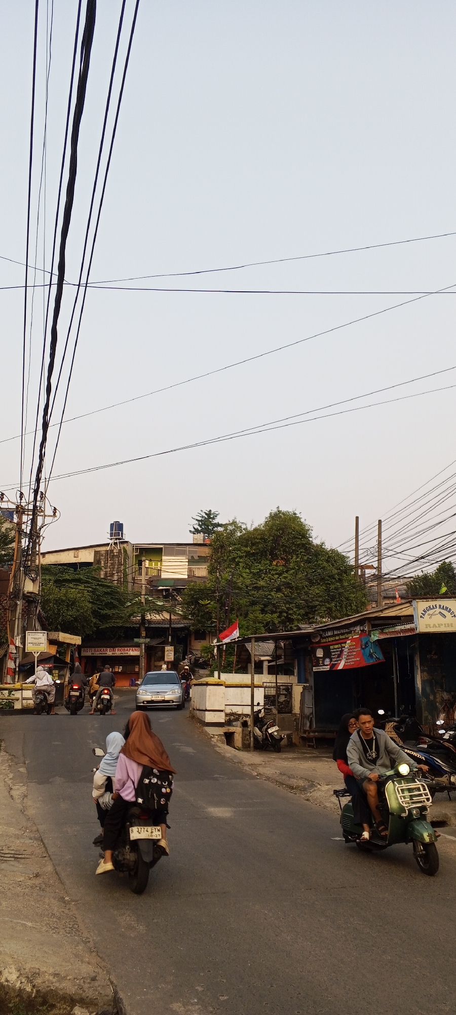 The image depicts a street scene in Depok, Indonesia, under a hazy sky. Several motorcycles are present, with two in the foreground carrying passengers. One motorcycle on the left has two individuals, a driver wearing a light blue hijab and a passenger in a pink shirt and brown hijab. On the right side of the street, a green scooter is ridden by a man wearing a gray jacket and a woman in a black hijab. A silver car is driving away from the viewer in the middle of the road. Buildings line the street, with one in the background appearing to be a multi-story structure with visible plumbing and electrical wiring. Businesses with awnings are also visible, one displaying a sign that reads "PANGKAS RAMBUT RAPIH" (Clean Haircut). Overhead, a dense network of power lines crisscrosses the sky. The overall impression is that of a typical urban street with ongoing traffic and commercial activity.