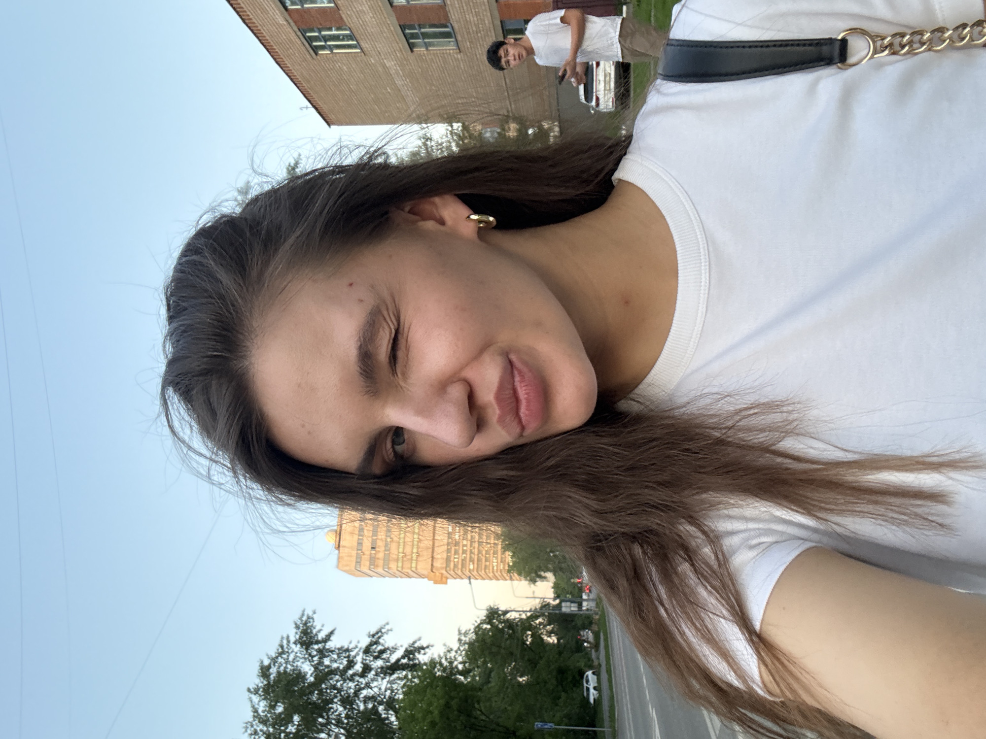 A young woman takes a selfie, winking at the camera. She has long brown hair and wears a white t-shirt. Behind her, a man wearing a white shirt and khaki pants stands near a parked white SUV, seemingly occupied with his phone. The setting appears to be a street in Moscow, Russia, judging from the architectural style of the buildings in the background. There are trees and traffic signals visible along the street. The sky is light blue.
