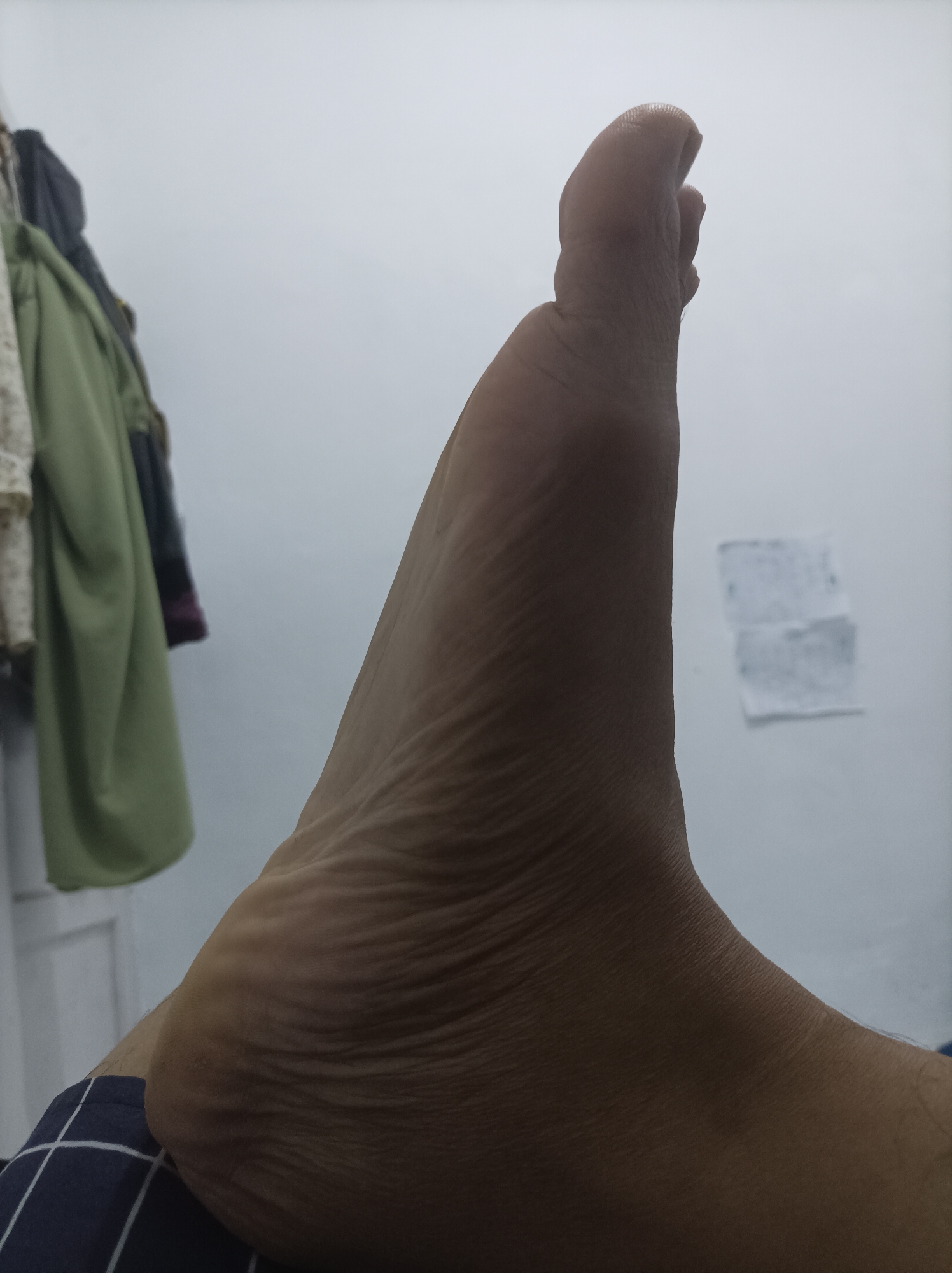 An overhead shot of a person's right foot, extended upwards with toes pointing to the right. The foot is positioned on a dark blue and white checkered surface. The background shows a white wall with some clothing hanging on the left side and a small, blurry piece of paper on the right. There is no indication of the location being in Pamengkang, Indonesia.