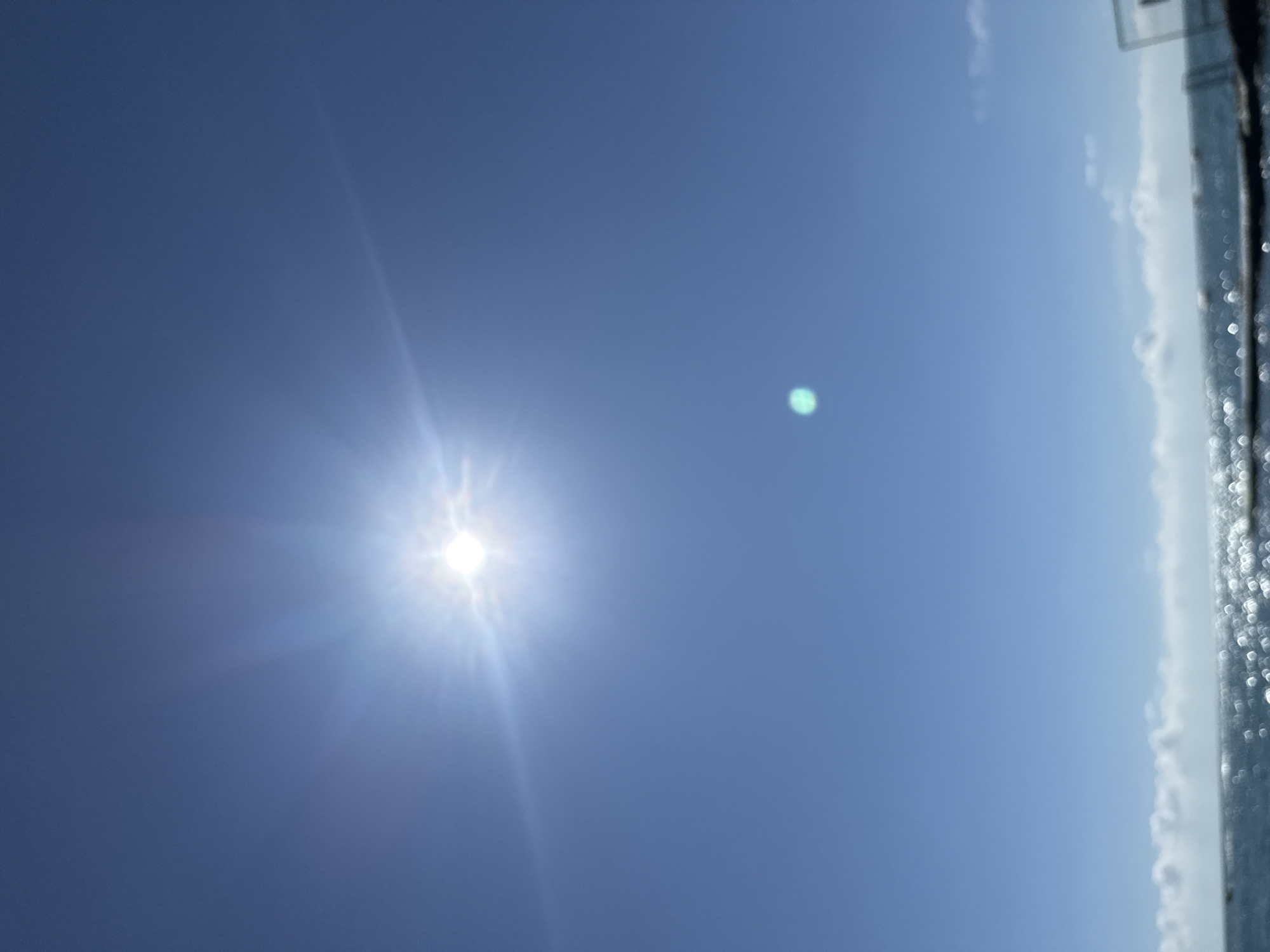 The image captures a bright, sunny day with a clear blue sky. The sun is prominently positioned in the upper left quadrant, emitting strong rays that create a lens flare effect. A smaller, out-of-focus green light artifact is visible in the upper right quadrant. The bottom of the image shows a body of water, likely the sea, with shimmering sunlight reflecting off its surface. A faint line of clouds is present near the horizon. The scene appears to be outdoors, possibly a coastal area. The presence of sunlight and the seascape suggests a location in Sochi, Russia, known for its Black Sea coastline and sunny weather, especially during warmer months. There are no visible people or specific structures that definitively place it within the city of Sochi, but the overall environment is consistent with its coastal geography.